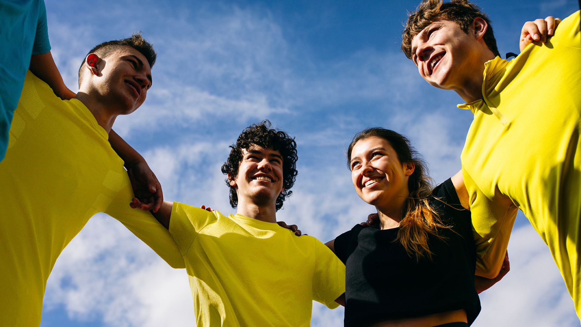 A group of young people with arms around each other in a circle, wearing yellow and smiling