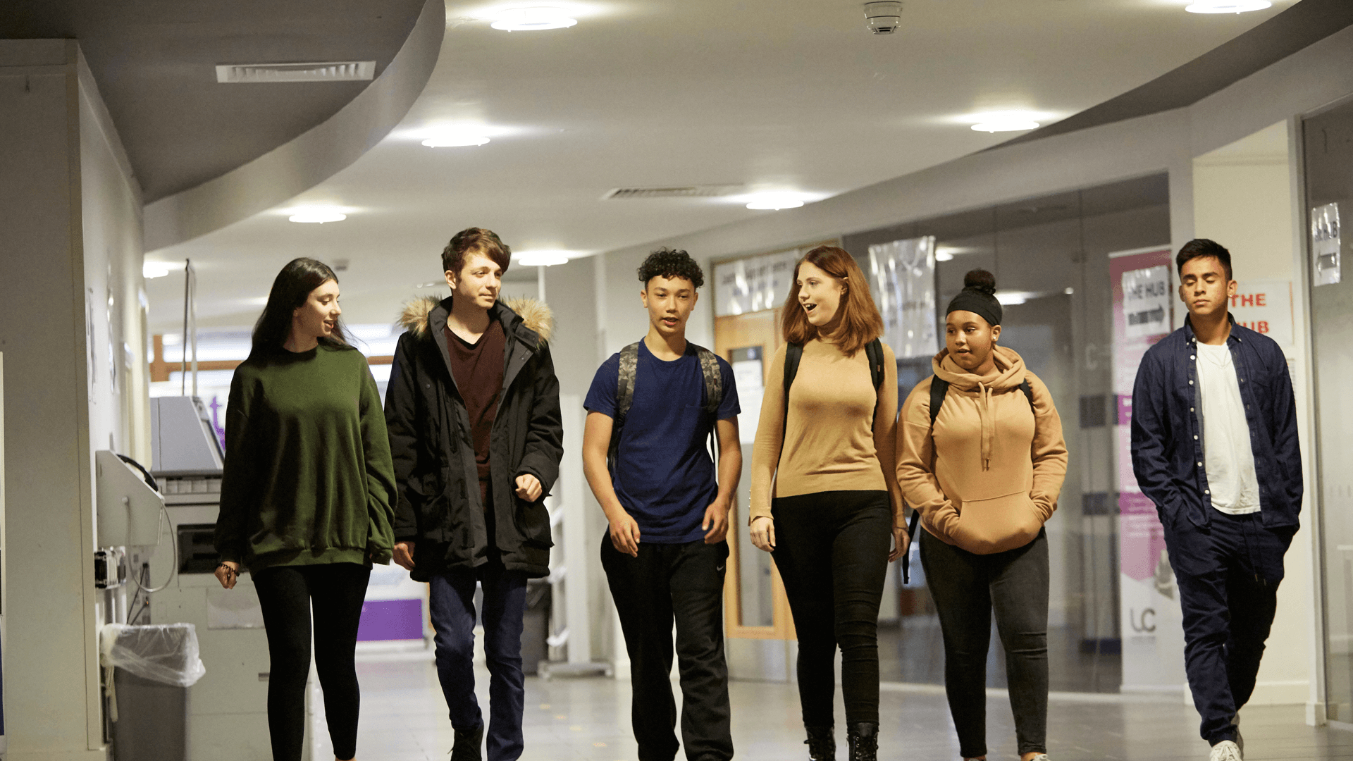 wide shot of six students walking and talking while walking in the hallway of the campus