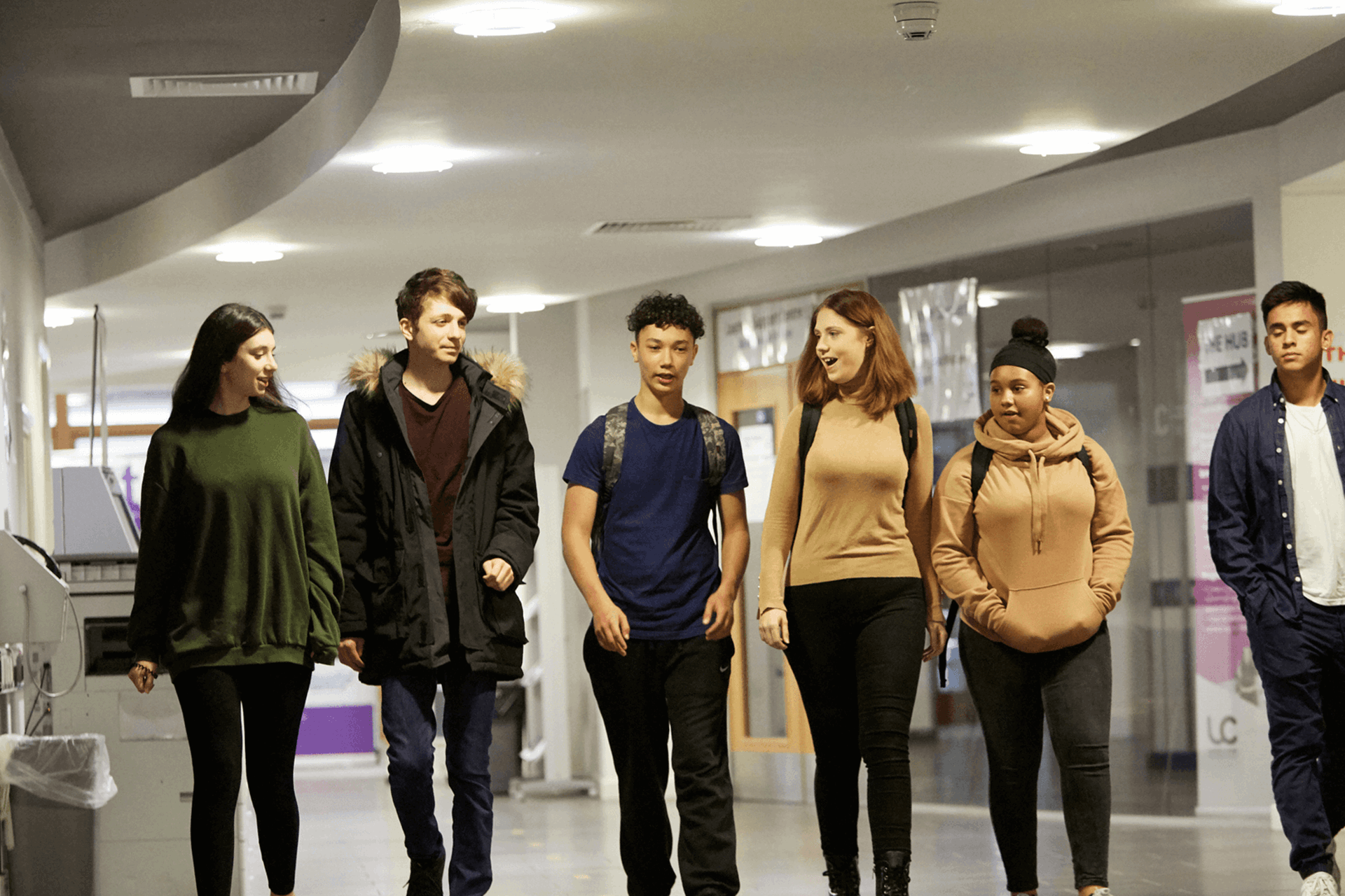 wide shot of six students walking and talking while walking in the hallway of the campus