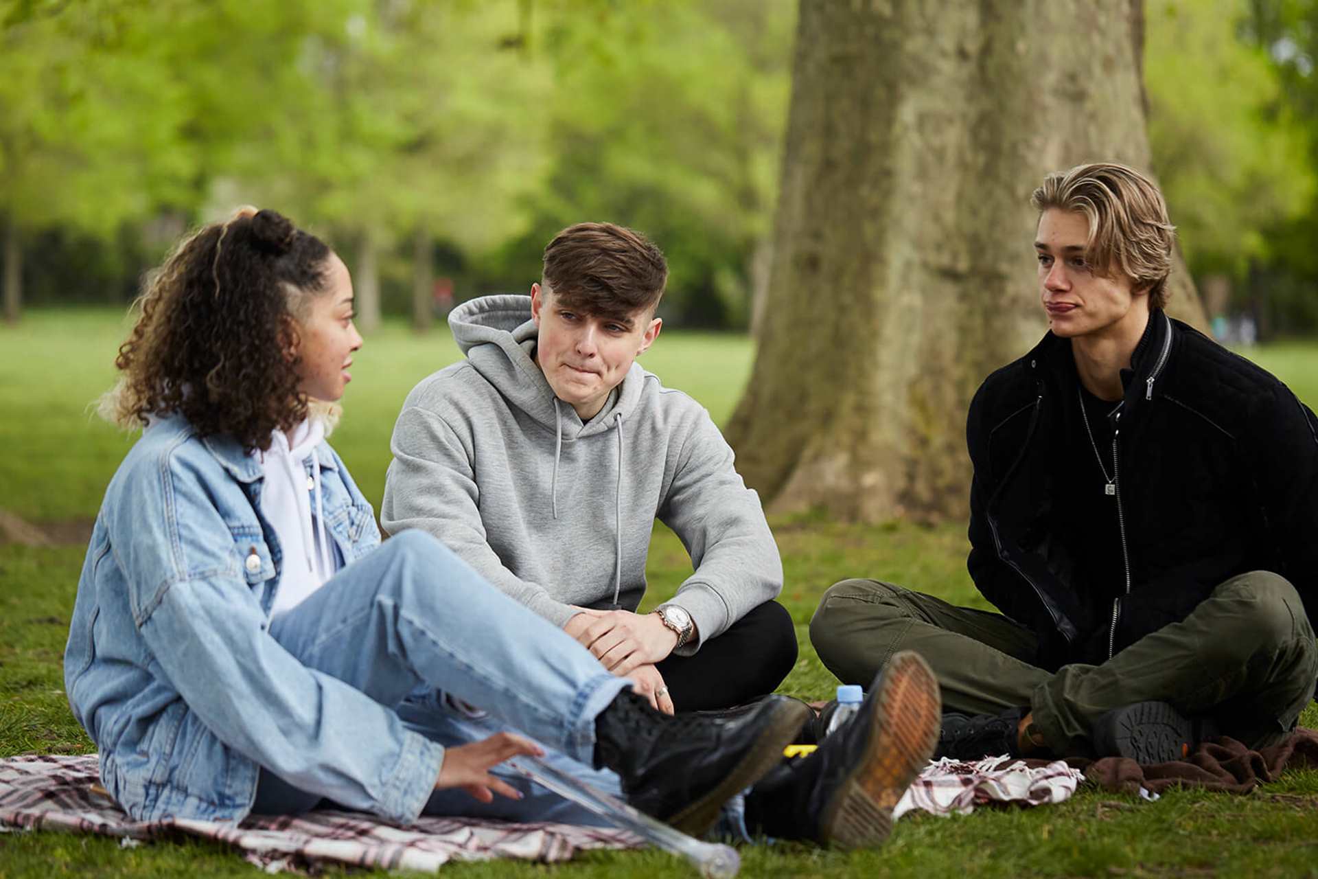 wide-shot-of-three-young-people-sitting-on-the-ground-together-talking-to-each-other-and-listening-with-trees-on-their-background