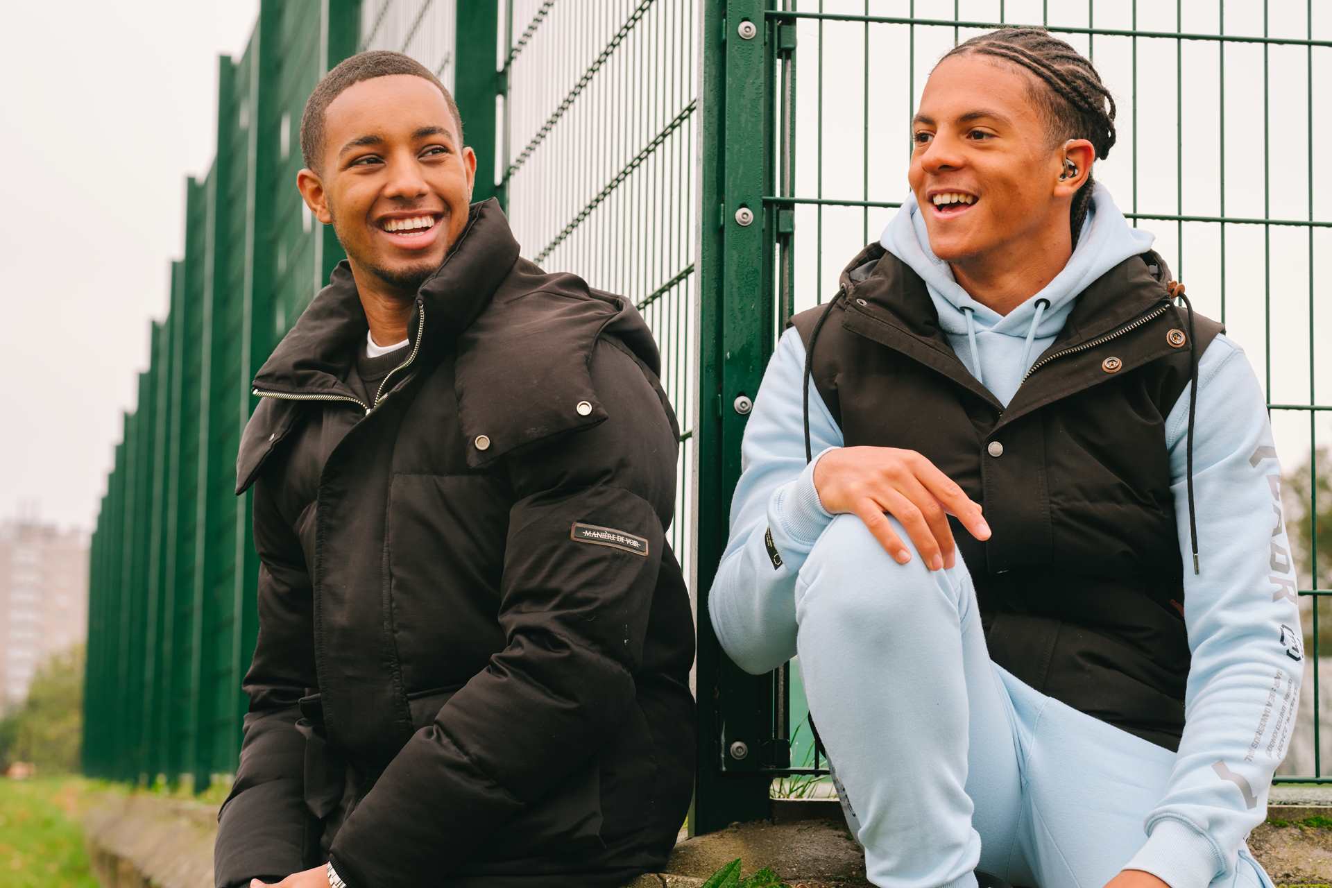 A young Black teenage boy wearing a hearing aid. He is laughing with a young Black man in the park.