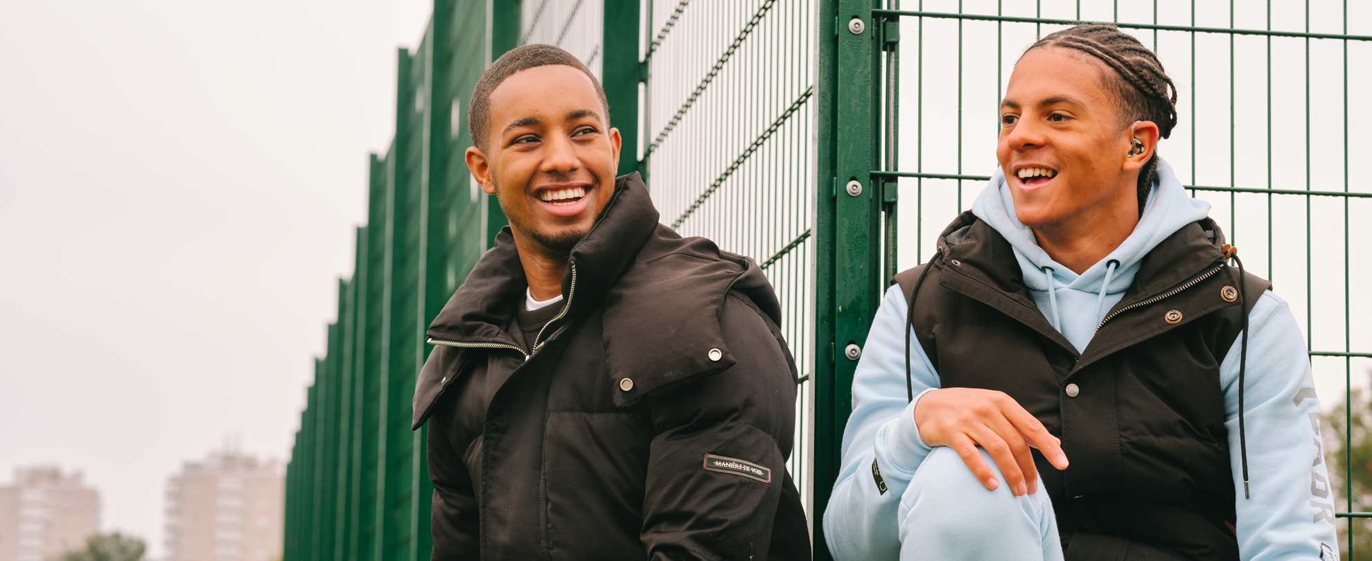 A young Black teenage boy wearing a hearing aid. He is laughing with a young Black man in the park.