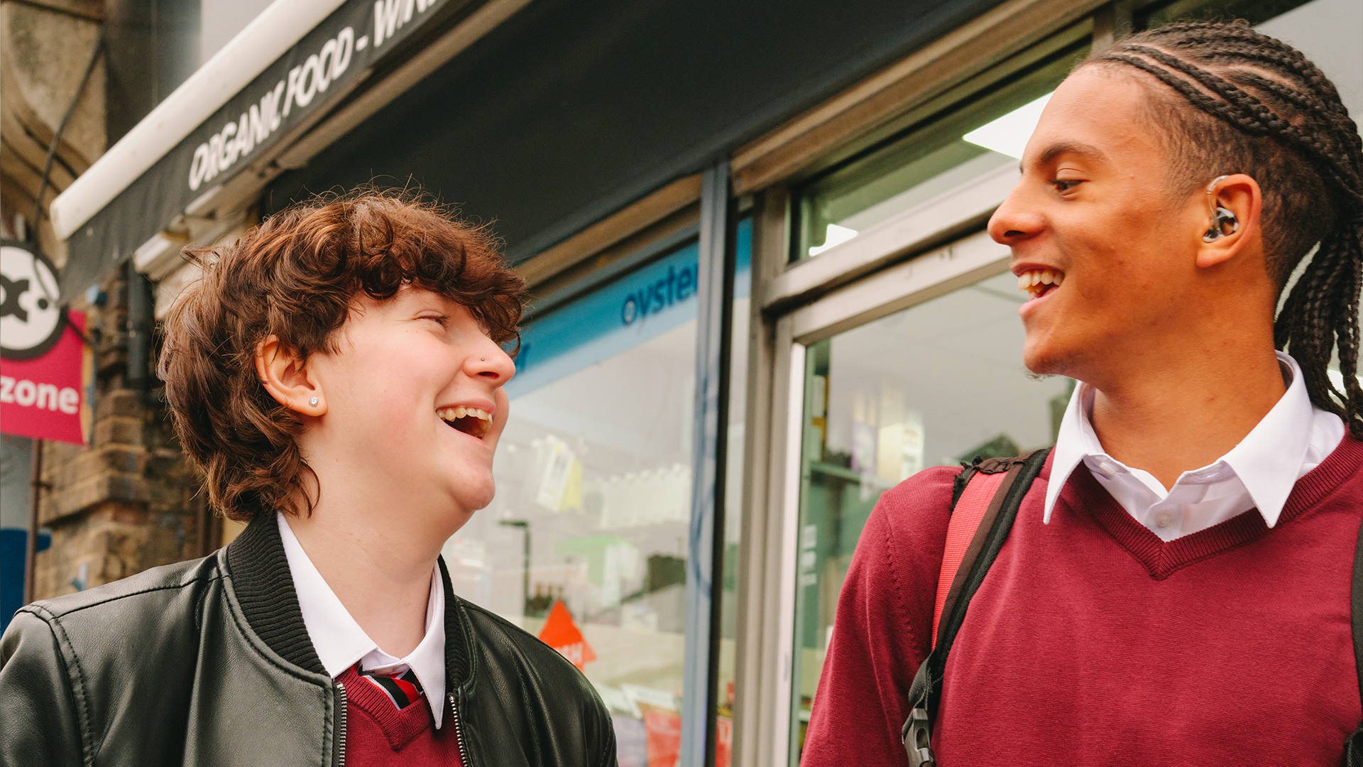 A Black teenage boy wearing a hearing aid laughing with a white non-binary teenager outside the shops.