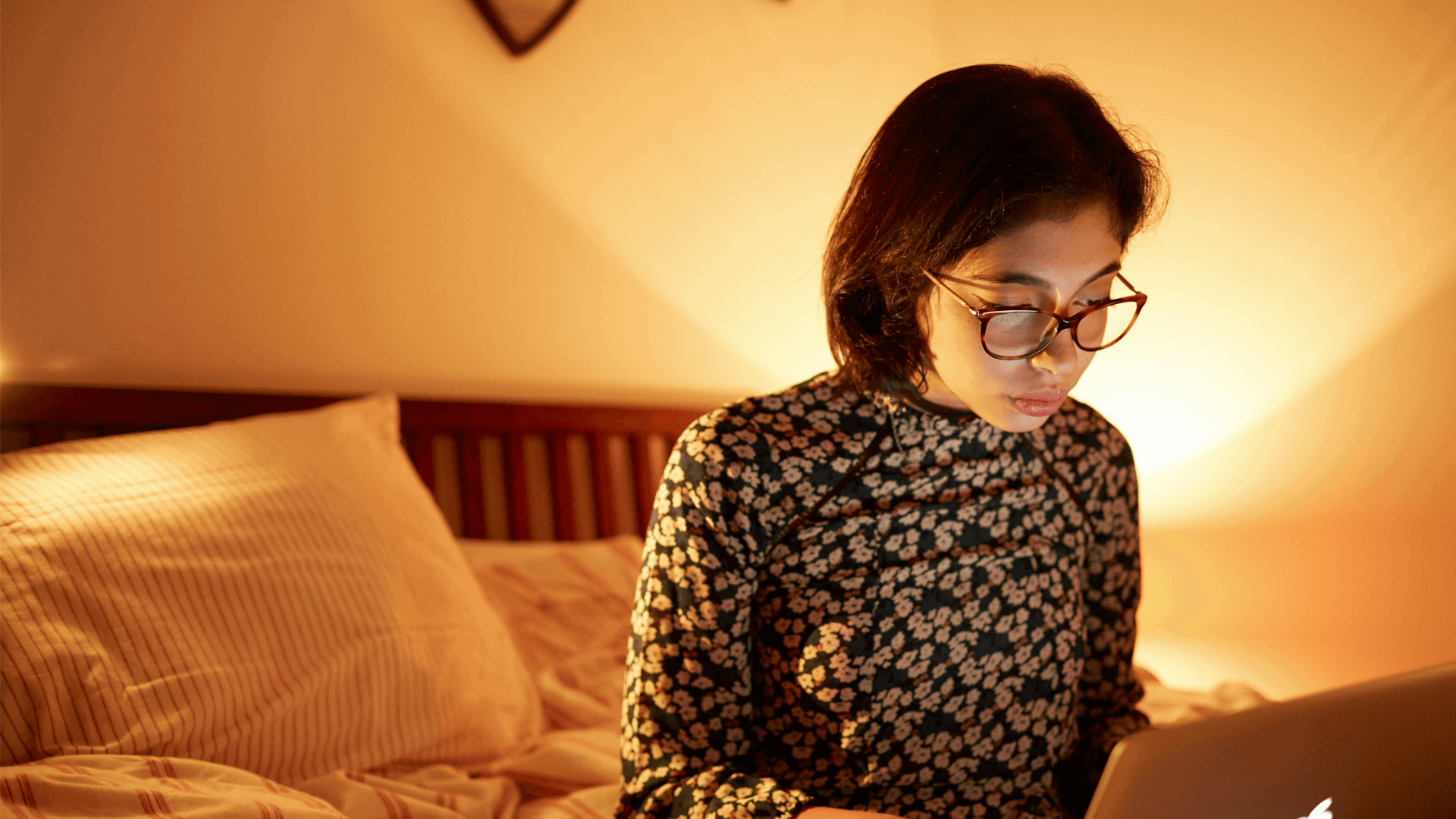 A girl wearing glasses sitting on the end of her bed while using her laptop.