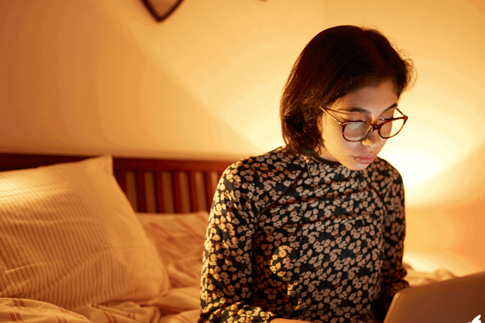 A girl wearing glasses sitting on the end of her bed while using her laptop.