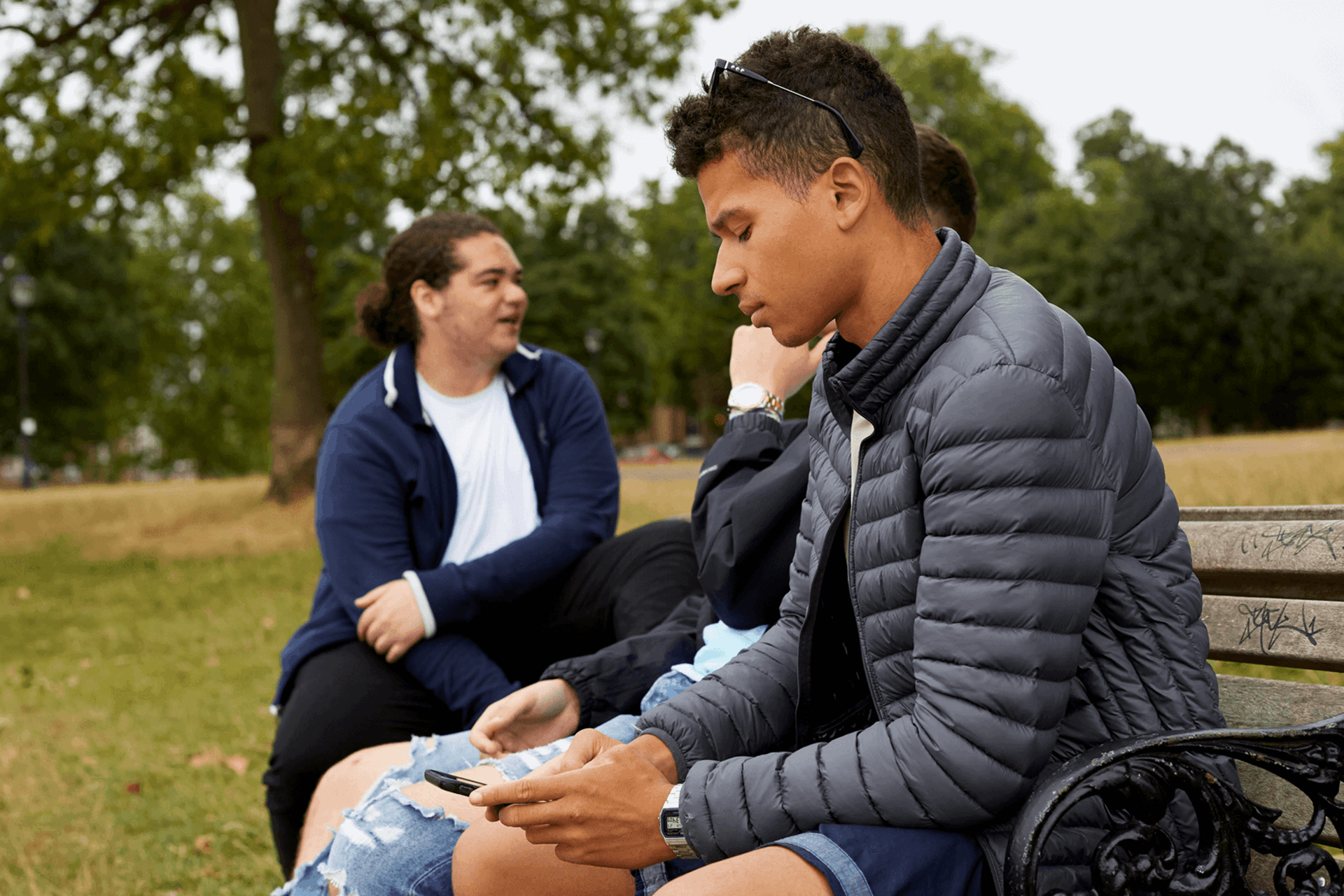 boy-in-a-jacket-looking-at-his-phone-ignoring-his-friends-who-are-talking-beside-him-while-sitting-on-a-bench-at-the-park