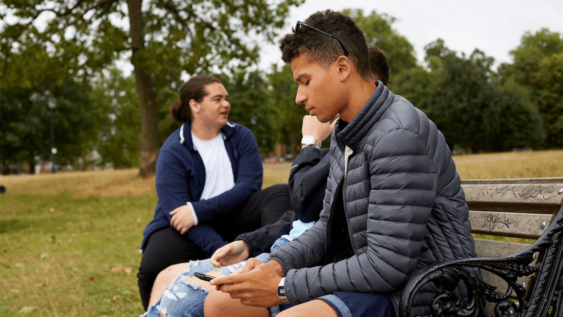 boy-in-a-jacket-looking-at-his-phone-ignoring-his-friends-who-are-talking-beside-him-while-sitting-on-a-bench-at-the-park