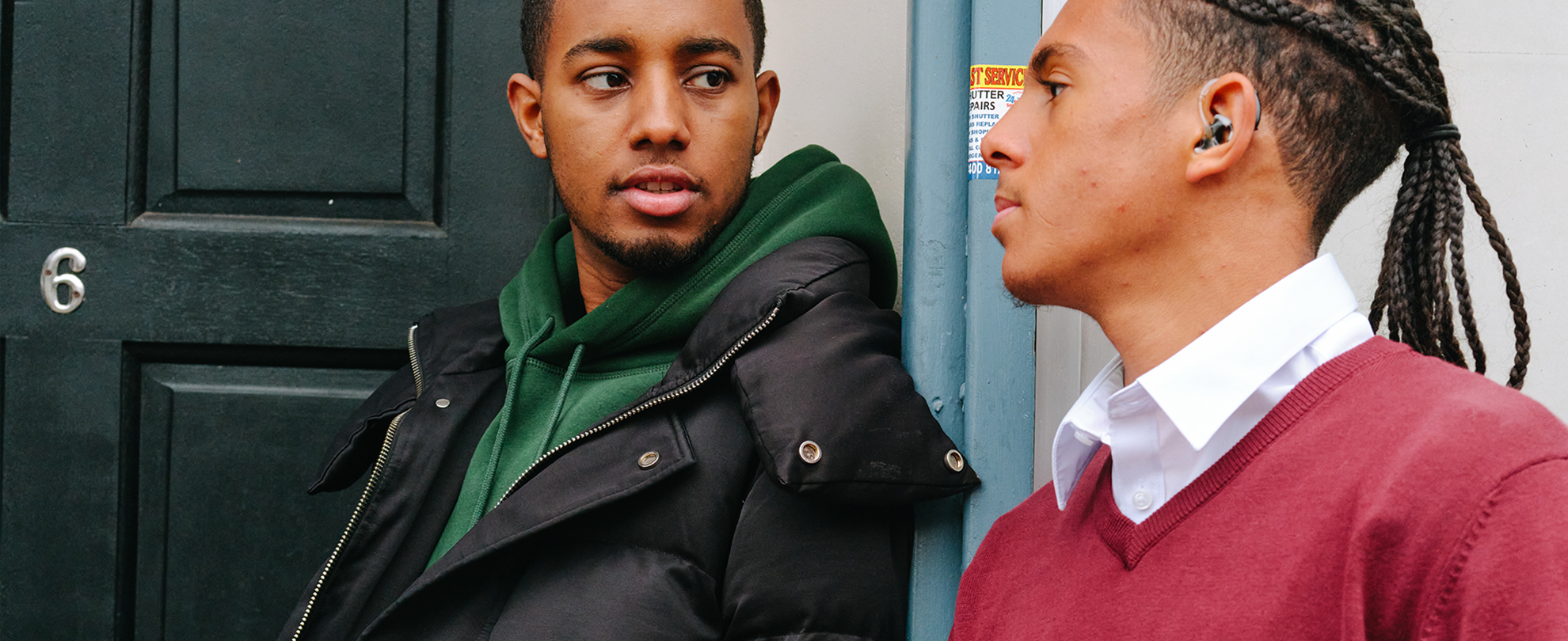 A young Black man standing outside a front door with a Black teenage boy wearing a hearing aid. They are talking together about something serious.