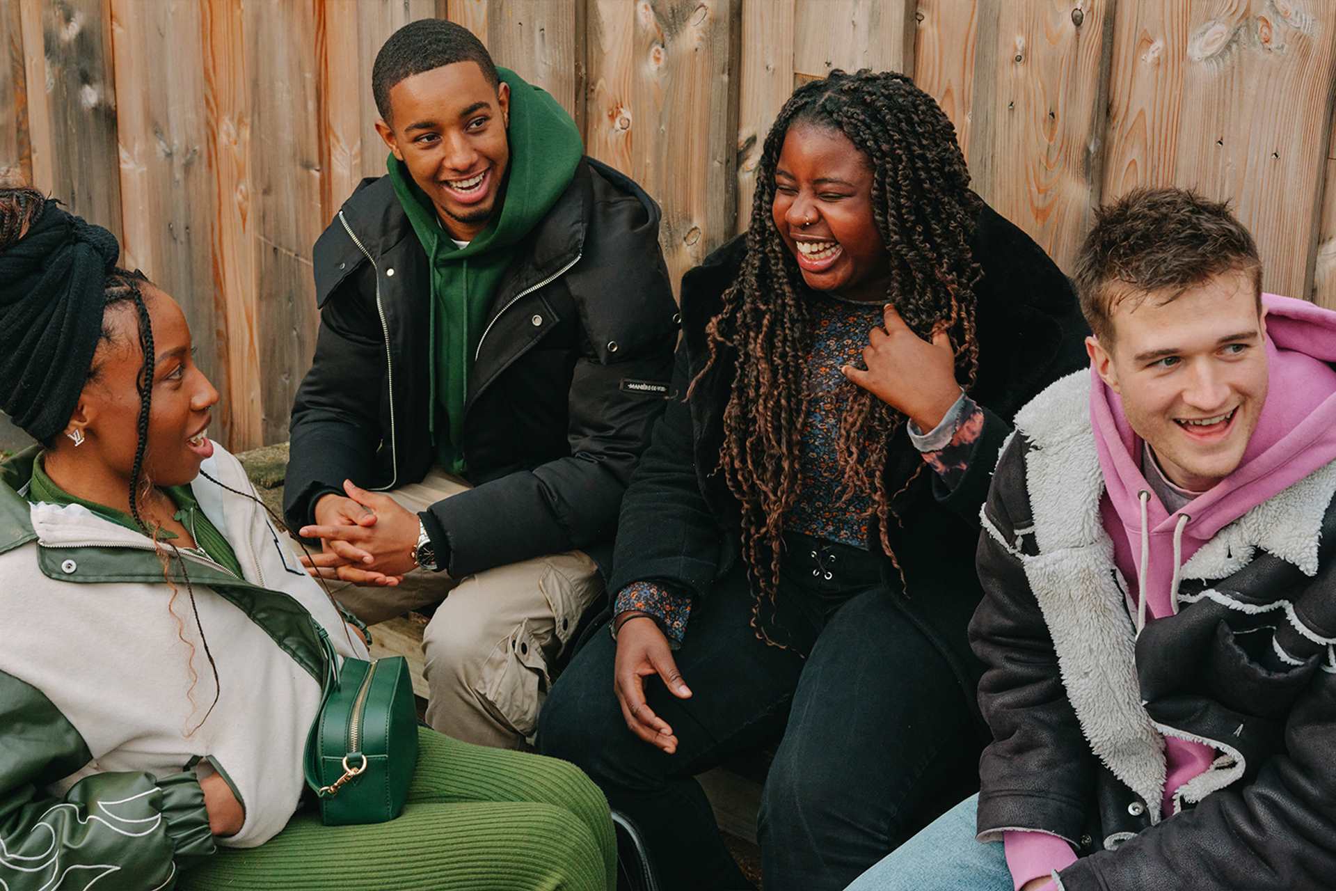 A group of young people laughing together outside on a bench. Group includes two Black young women (one in a wheelchair), one Black young man, and a white young man.