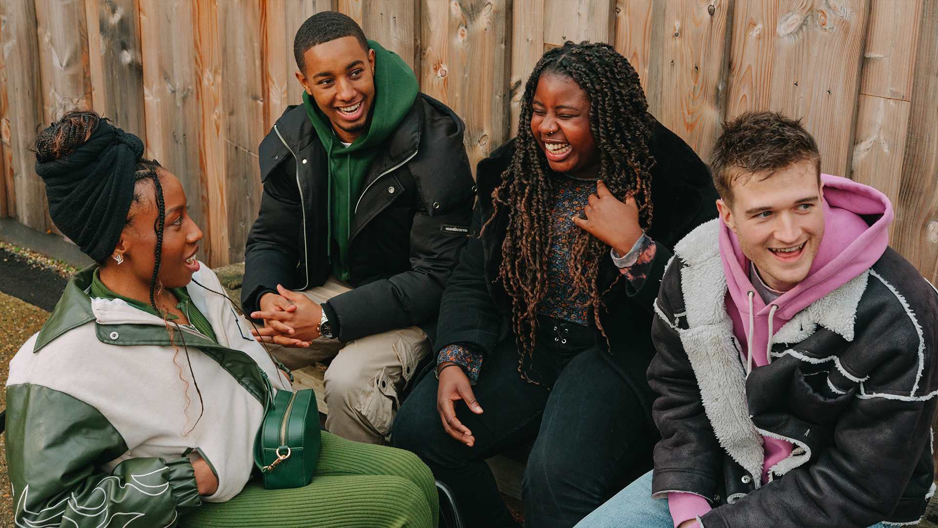 A group of young people laughing together outside on a bench. Group includes two Black young women (one in a wheelchair), one Black young man, and a white young man.