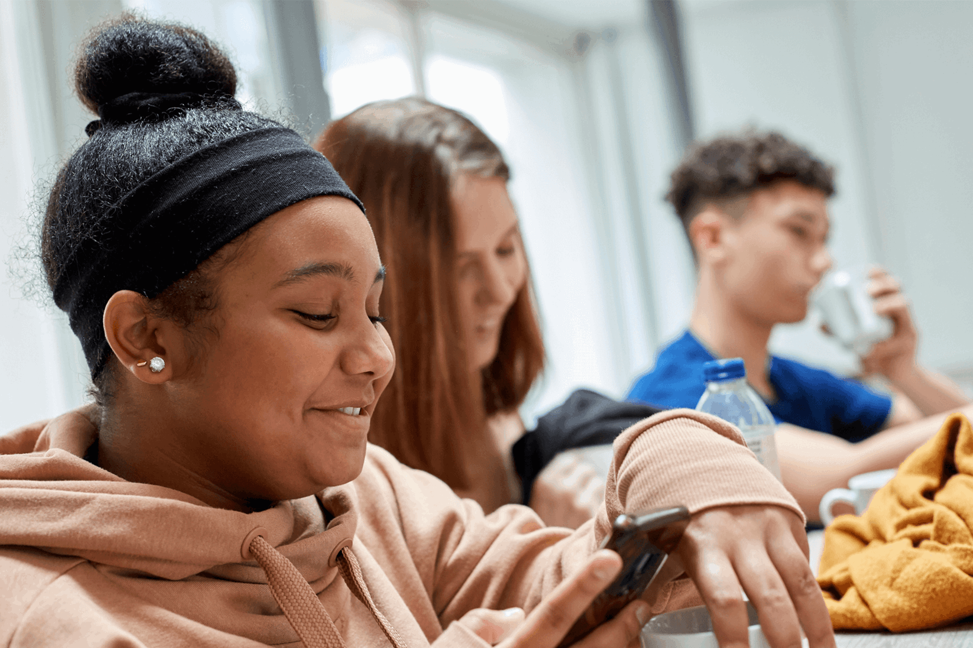 Three young people sitting together in a classroom. They are smiling while looking at their mobile phones.