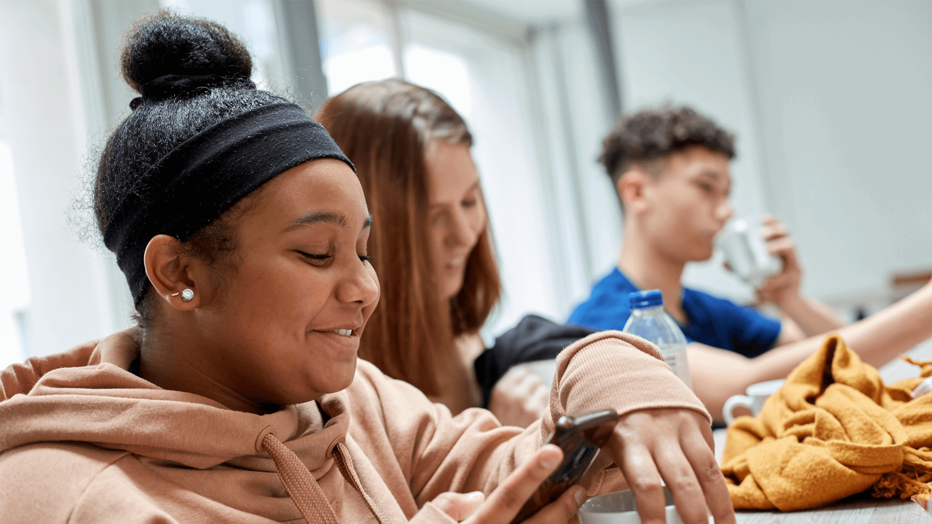 Three young people sitting together in a classroom. They are smiling while looking at their mobile phones.