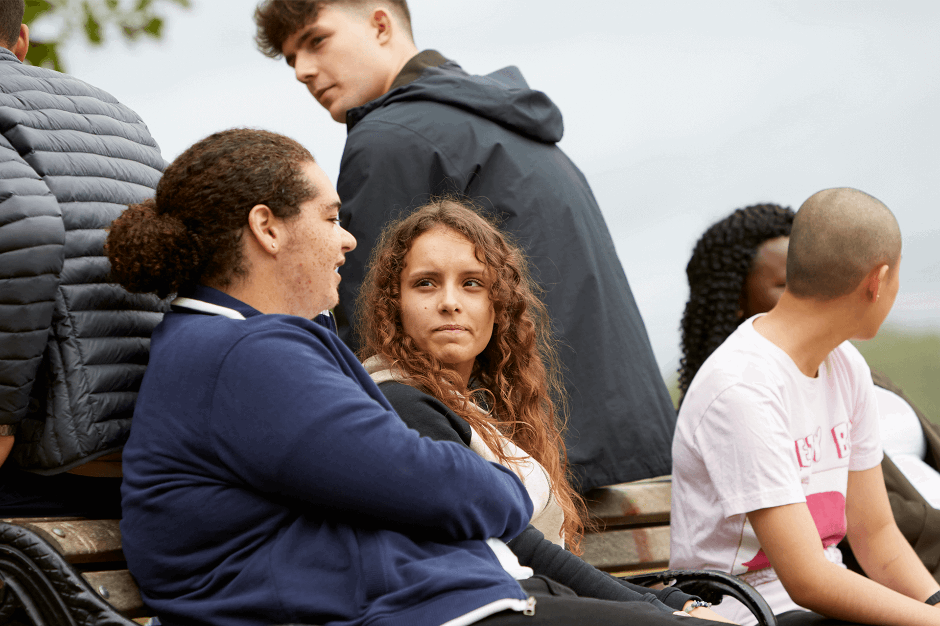 group of six young people talking and listening to one another while sitting on a bench in a park