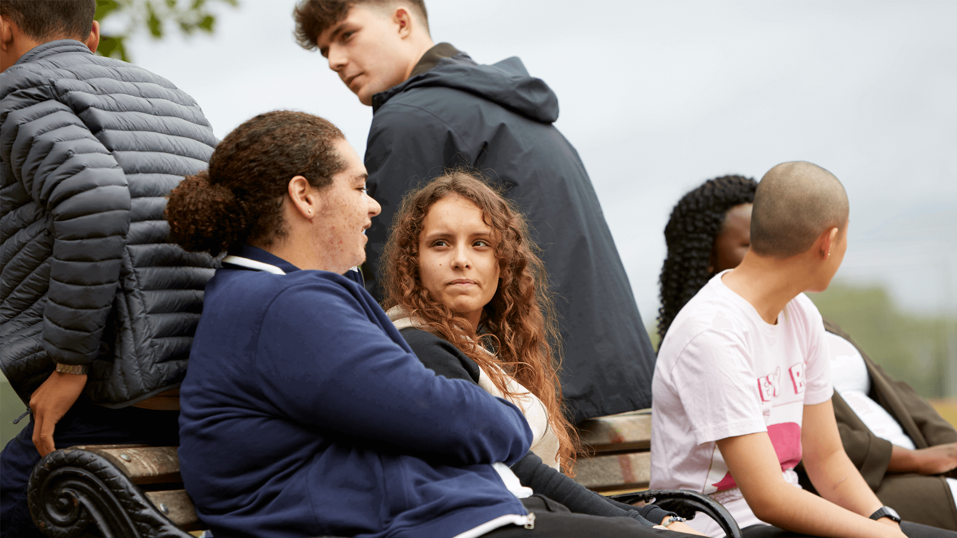 group of six young people talking and listening to one another while sitting on a bench in a park