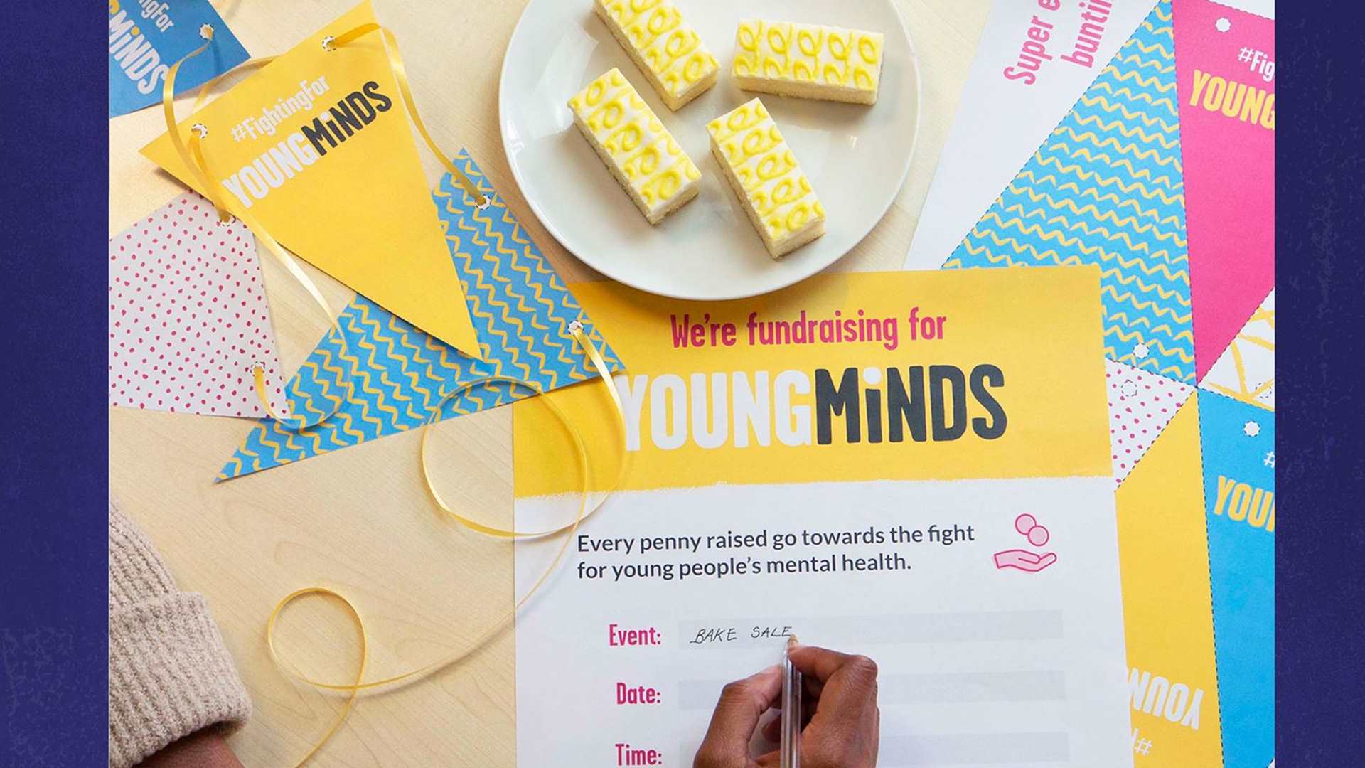 YoungMinds bunting is placed all over a desk with yellow ribbon. There are some yellow cake slices and our fundraising event poster which someone is writing on.