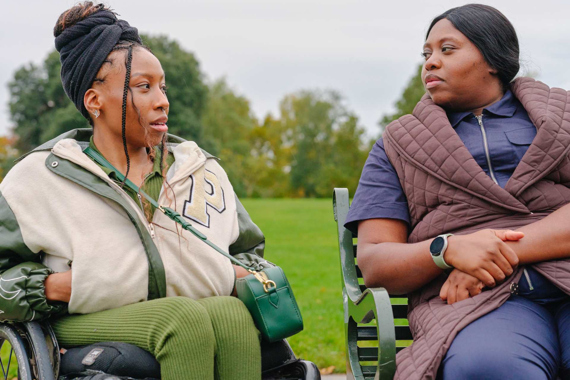 A young Black woman in a wheelchair and an older Black woman sitting on a bench in the park. They are talking about something serious.