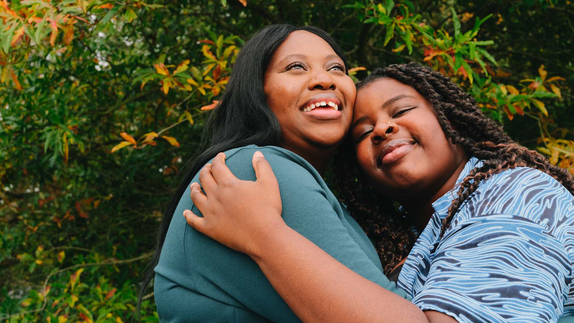 A young Black woman hugging an older Black woman in the park on a bench. They are both smiling.