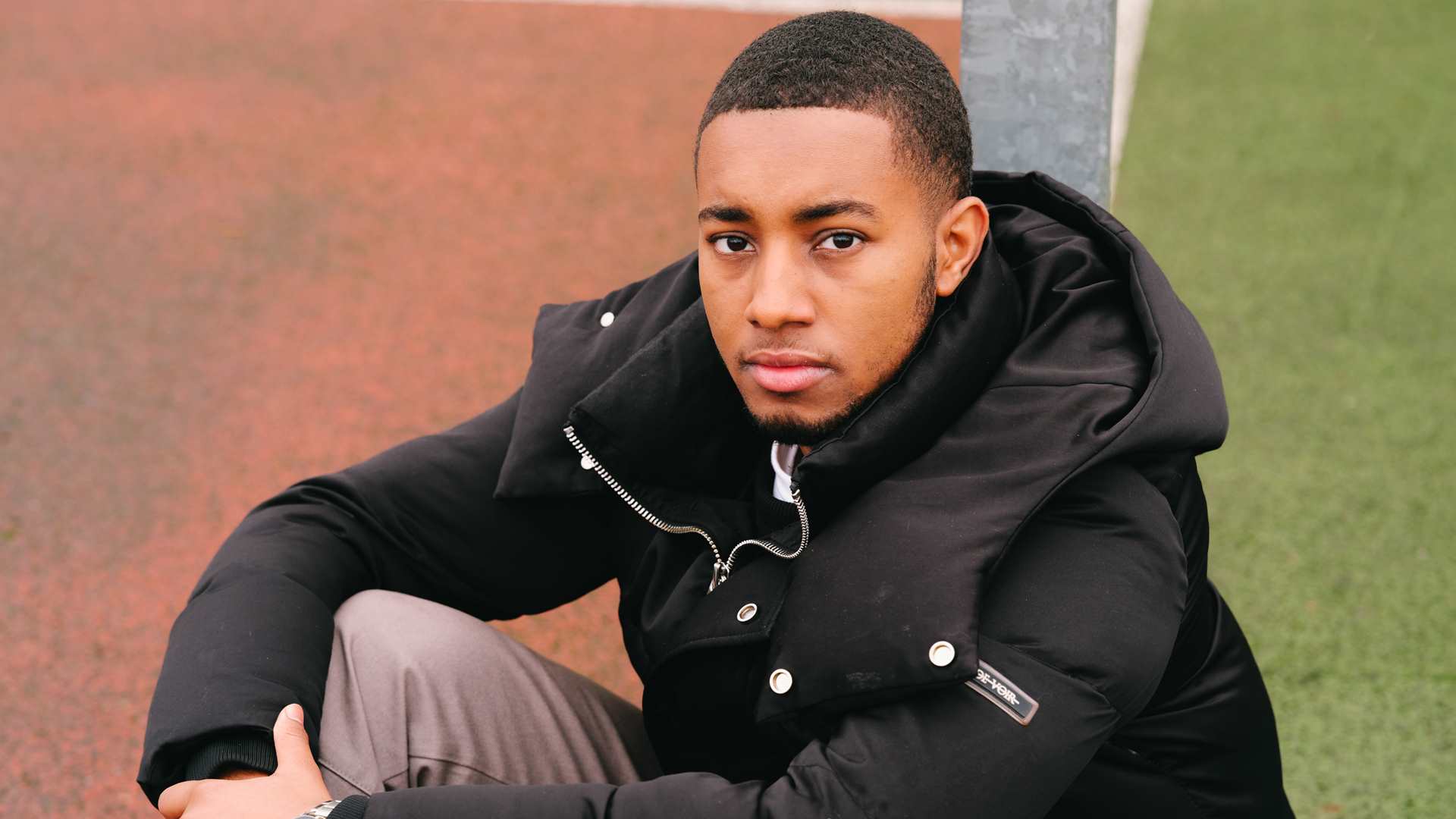 A young Black man sitting on the ground in the park and staring into the camera.