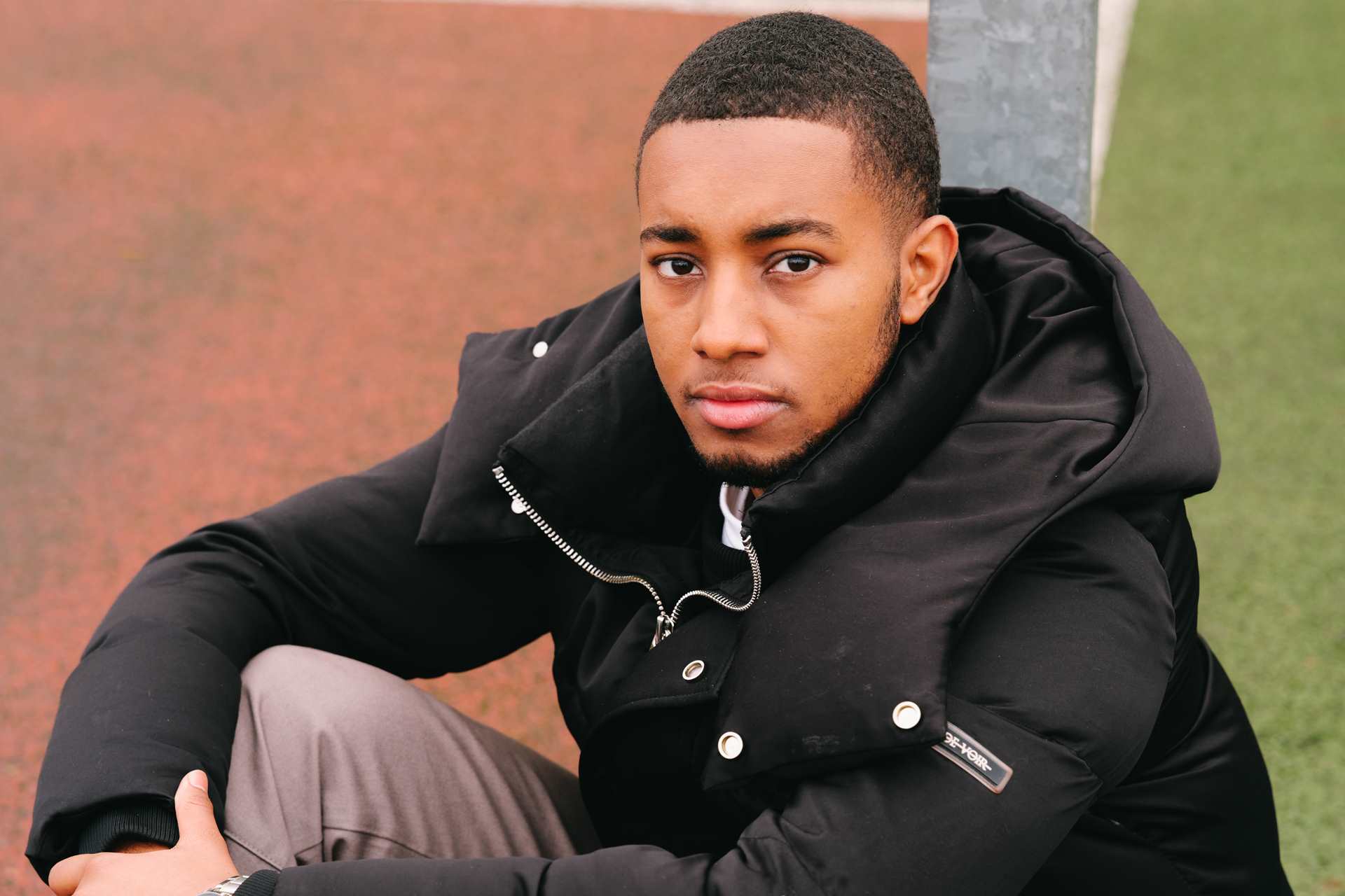 A young Black man sitting on the ground in the park and staring into the camera.
