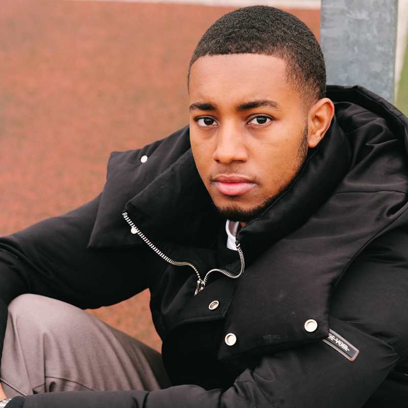 A young Black man sitting on the ground in the park and staring into the camera.
