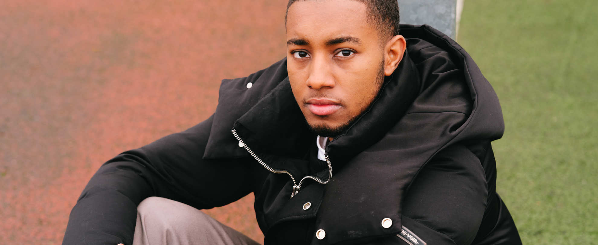 A young Black man sitting on the ground in the park and staring into the camera.