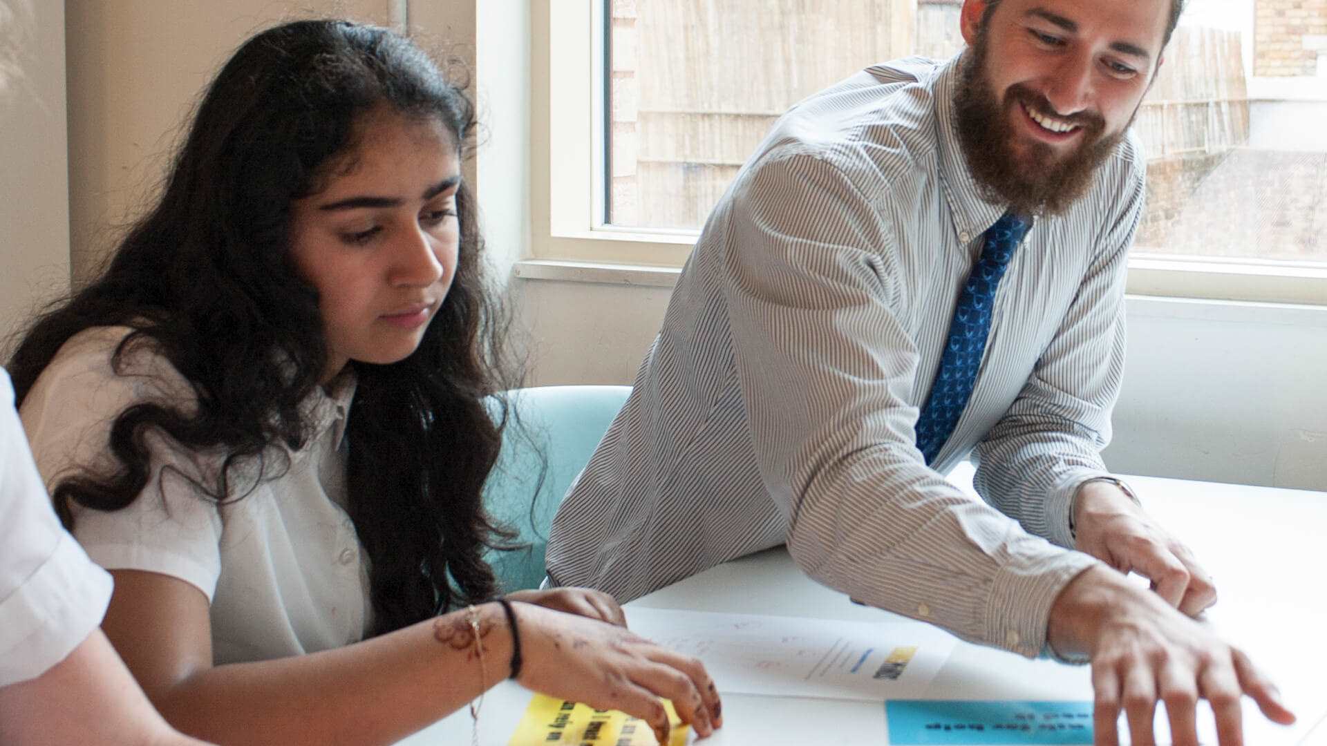 a teacher smiling while he sits beside a student to help her work in an activity