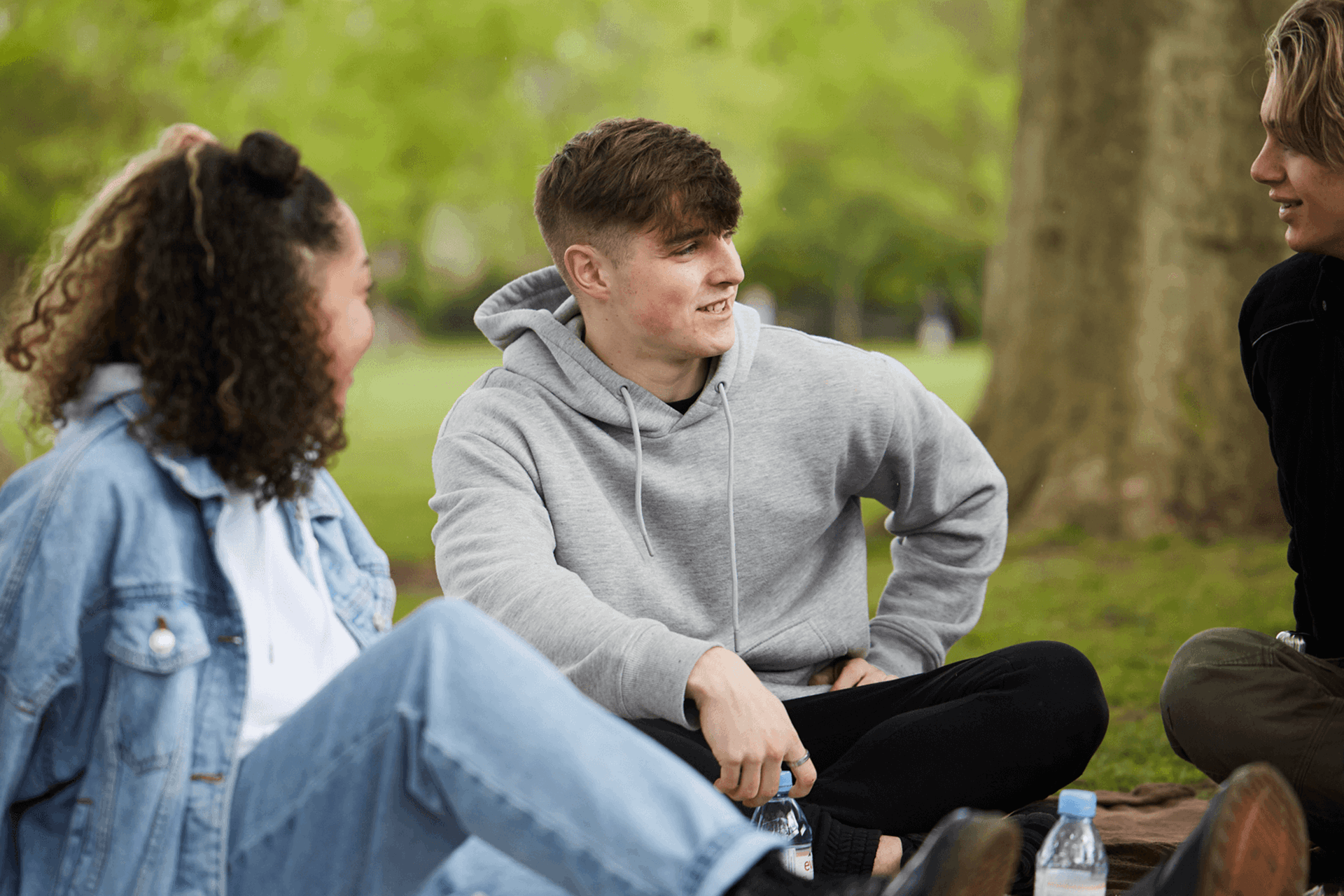 Three young people talking while sitting in the grass in a park.