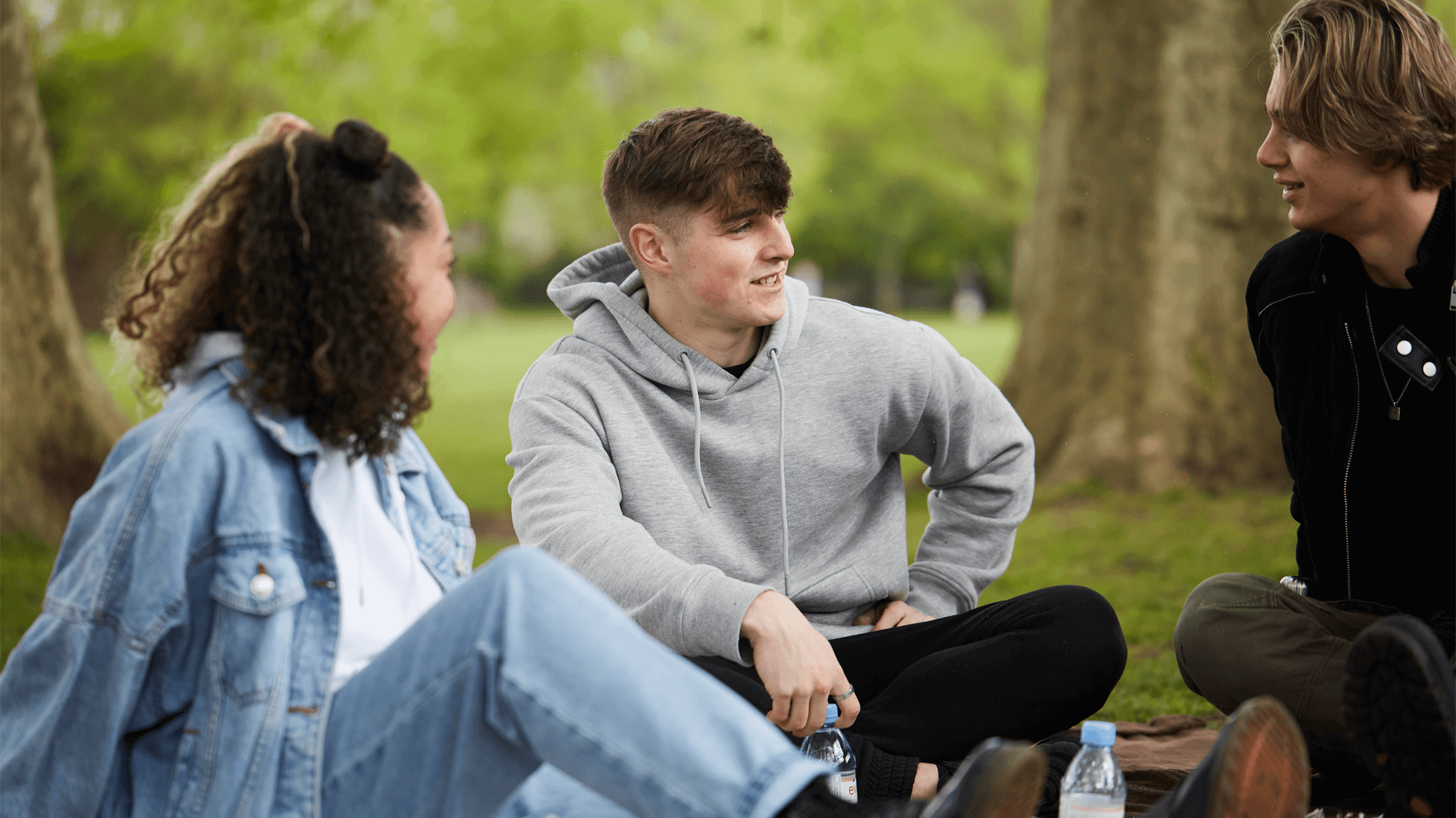 Three young people talking while sitting in the grass in a park.