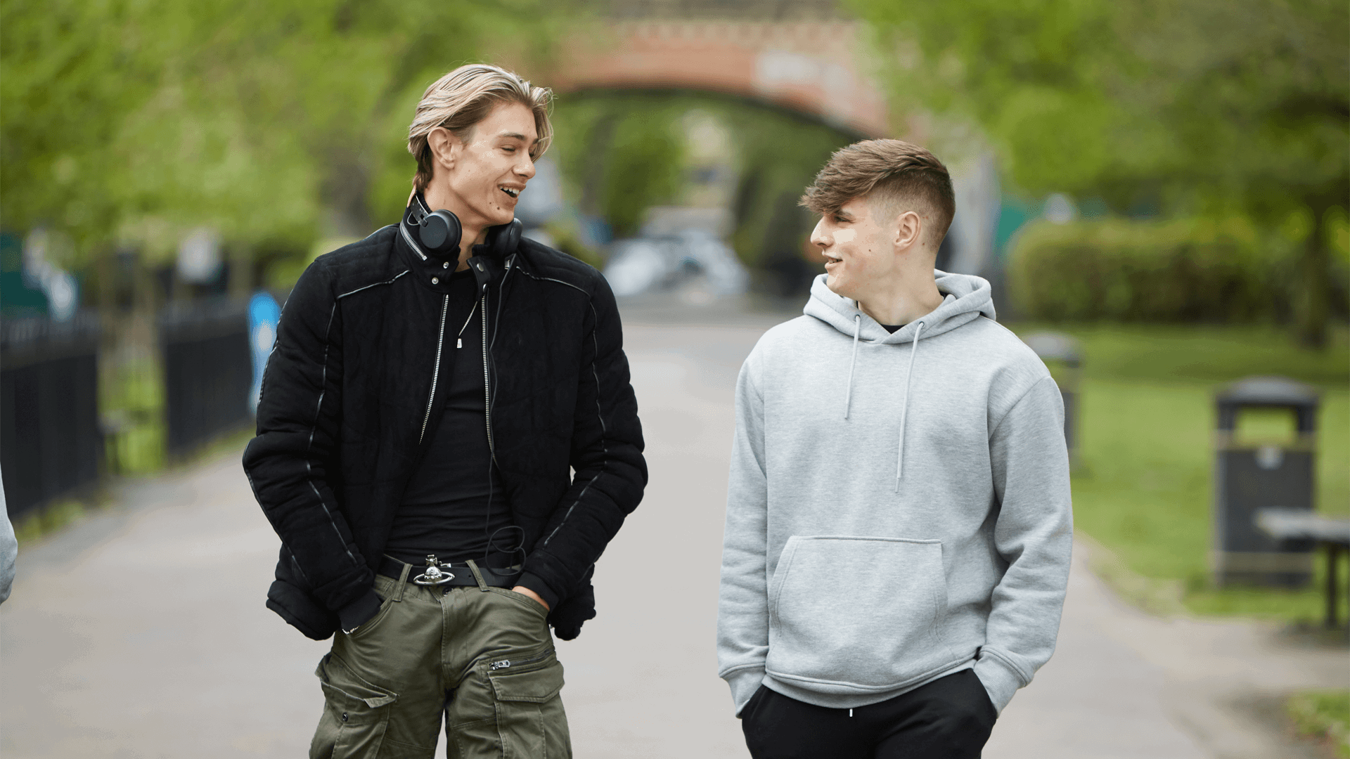 Two young men walking down a together, talking and smiling. One is wearing a black jacket and headphones around his neck. The other is wearing a grey hoodie.