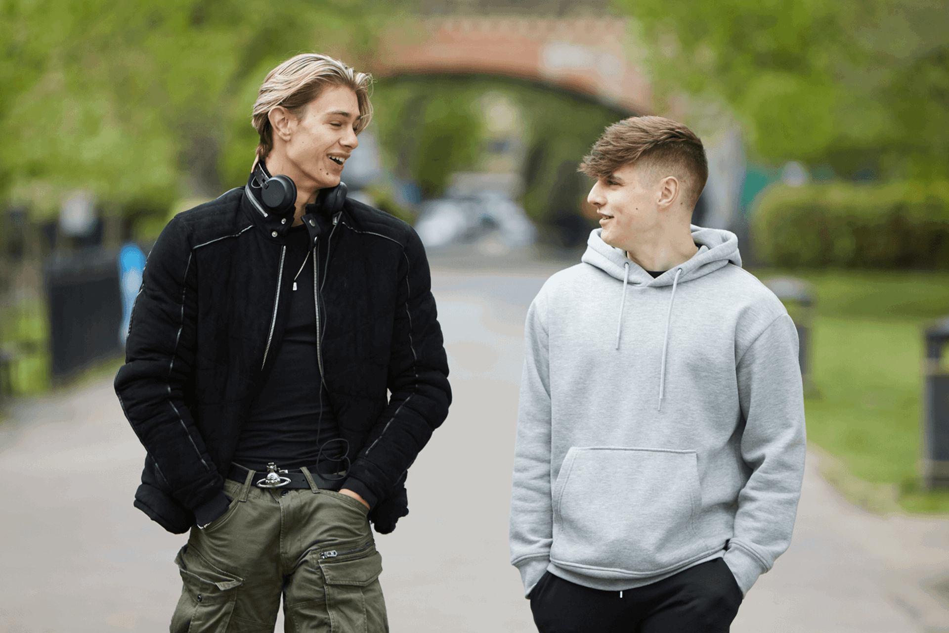 Two young men walking down a together, talking and smiling. One is wearing a black jacket and headphones around his neck. The other is wearing a grey hoodie.