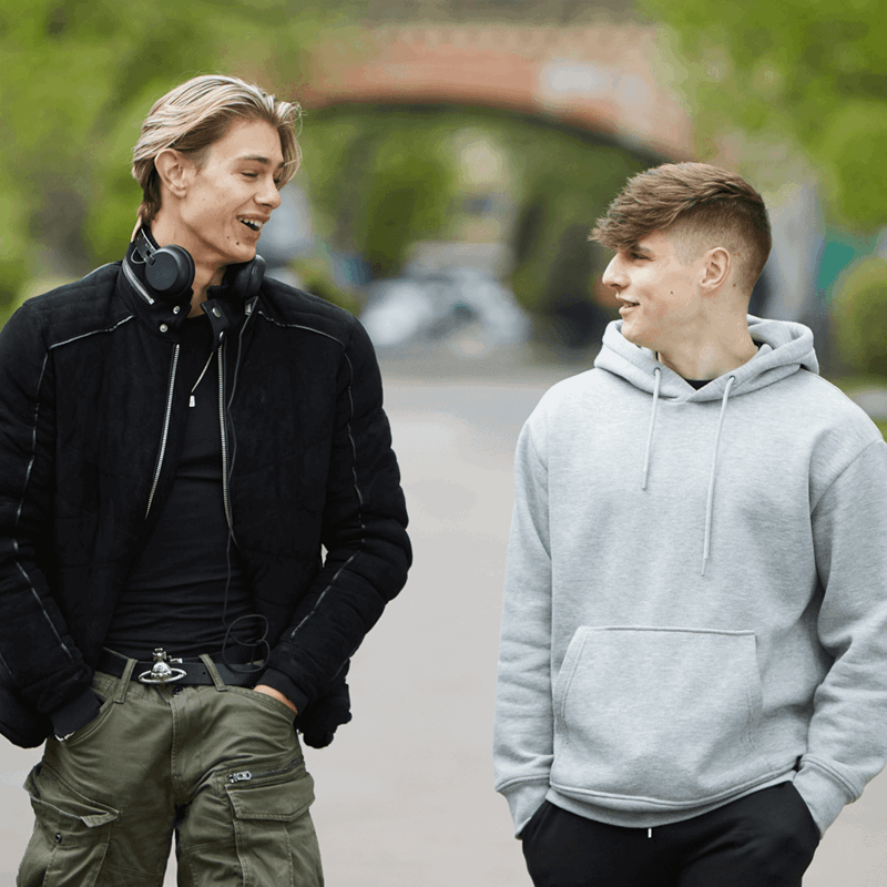 two-young-man-wearing-black-jacket-with-headphones-on-his-neck-and-hands-on-the-pockets-of-his-trousers-talking-while-walking-with-another-boy-wearin-grey-hoodie-on-a-street