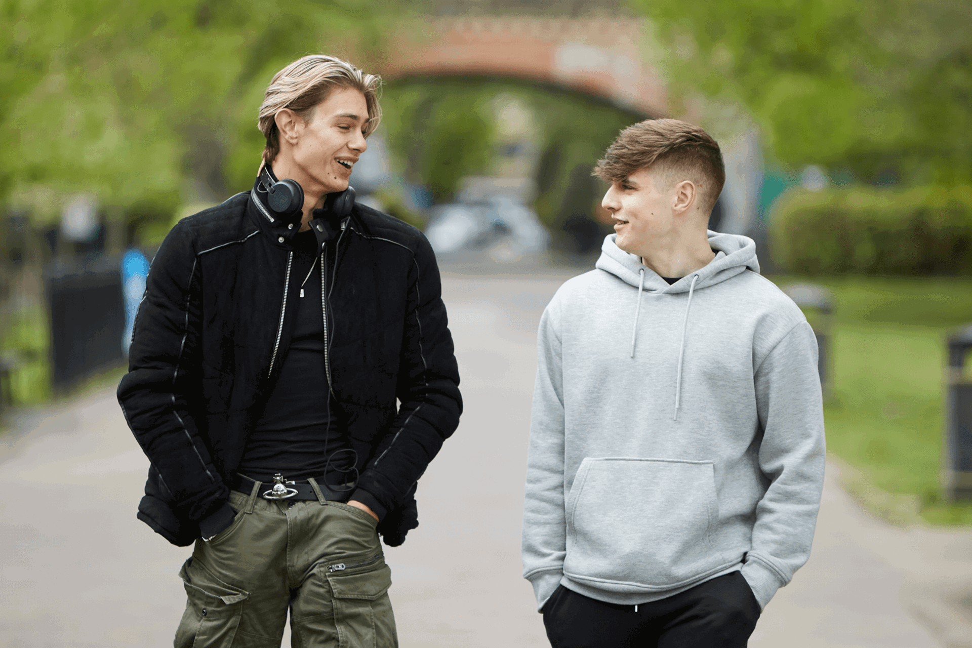 Two young men walking down a together, talking and smiling. One is wearing a black jacket and headphones around his neck. The other is wearing a grey hoodie.