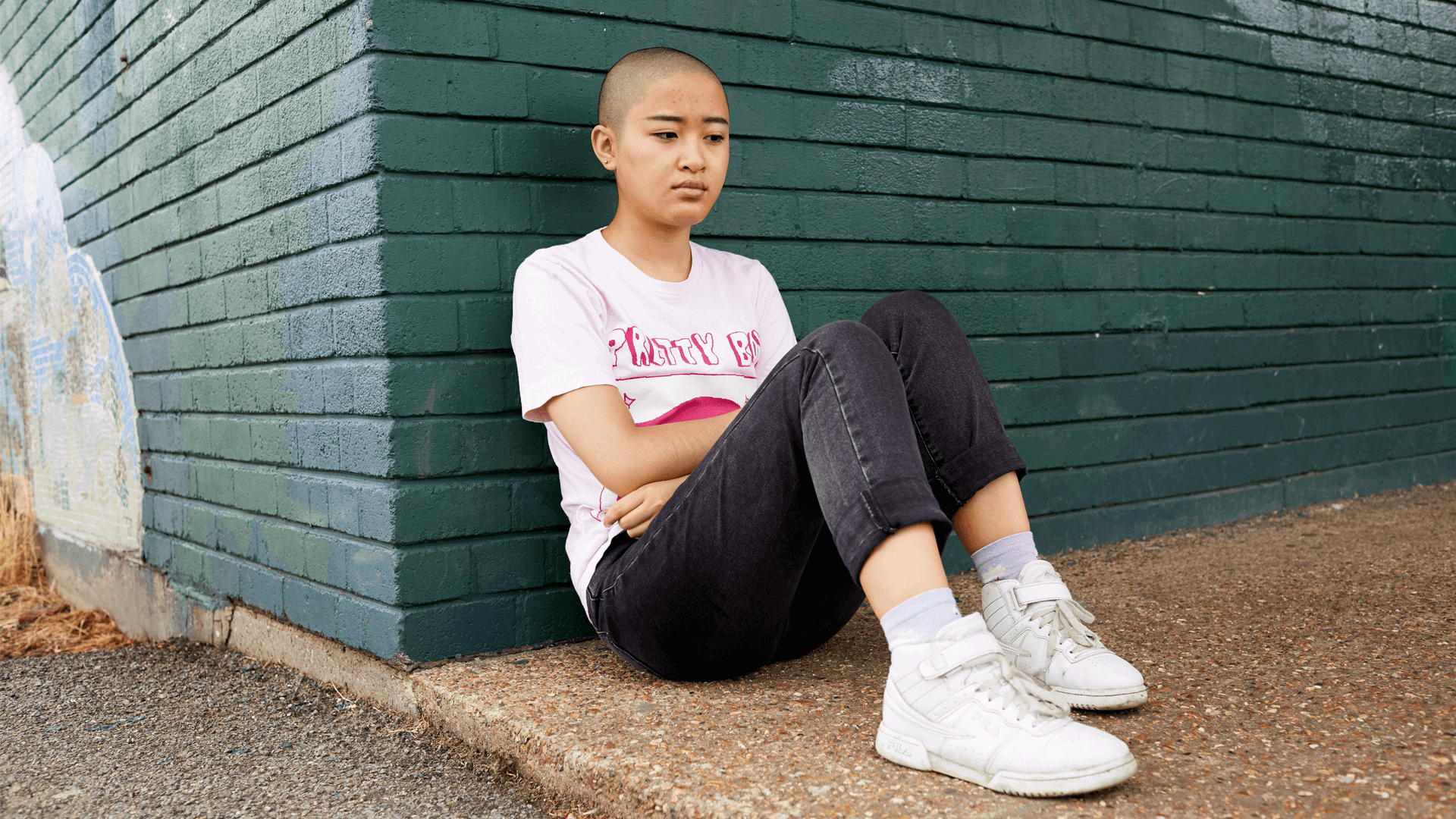 A girl with a shaved head wearing a t-shirt and black jeans. She is sitting on the ground and leaning against a green brick wall.