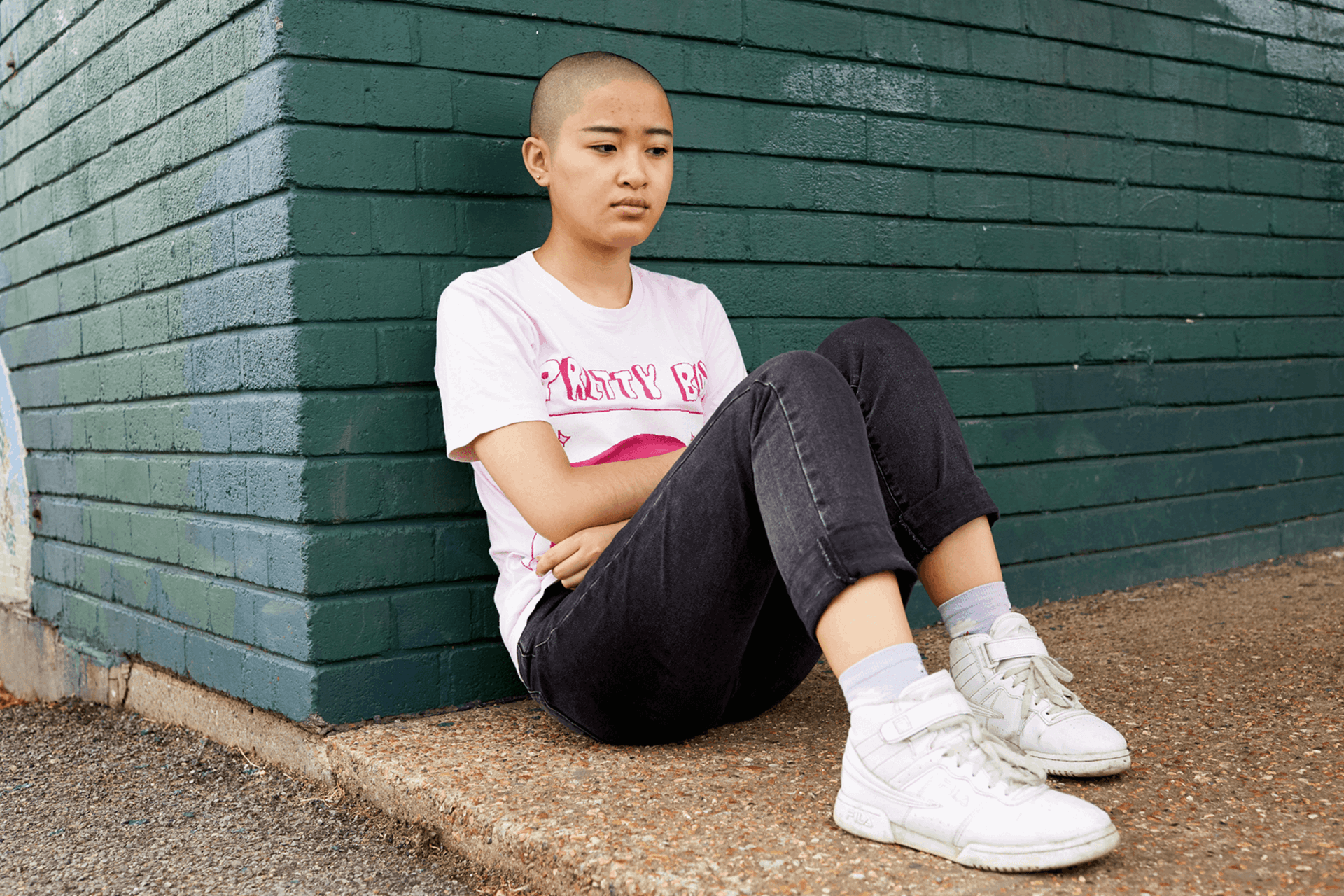 A girl with a shaved head wearing a t-shirt and black jeans. She is sitting on the ground and leaning against a green brick wall.