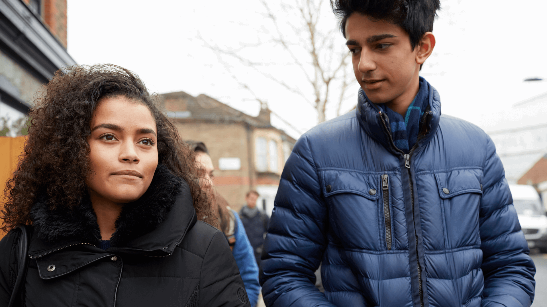 Two young people wearing coats and walking together down a street.