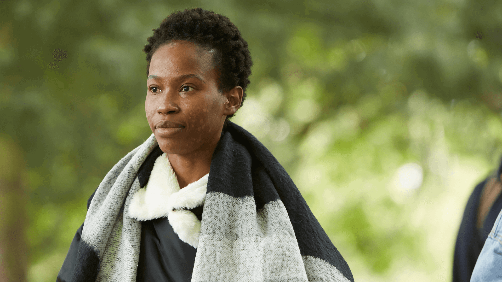 A young woman wearing a grey and black scarf walking through a park.