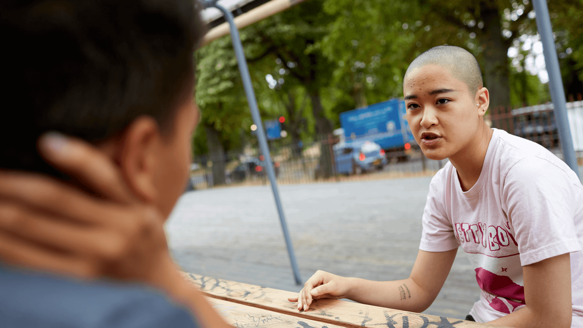 girl with a shaved head wearing white shirt explaining to a friend in front of her while sitting on a dining bench with a busy street on the background