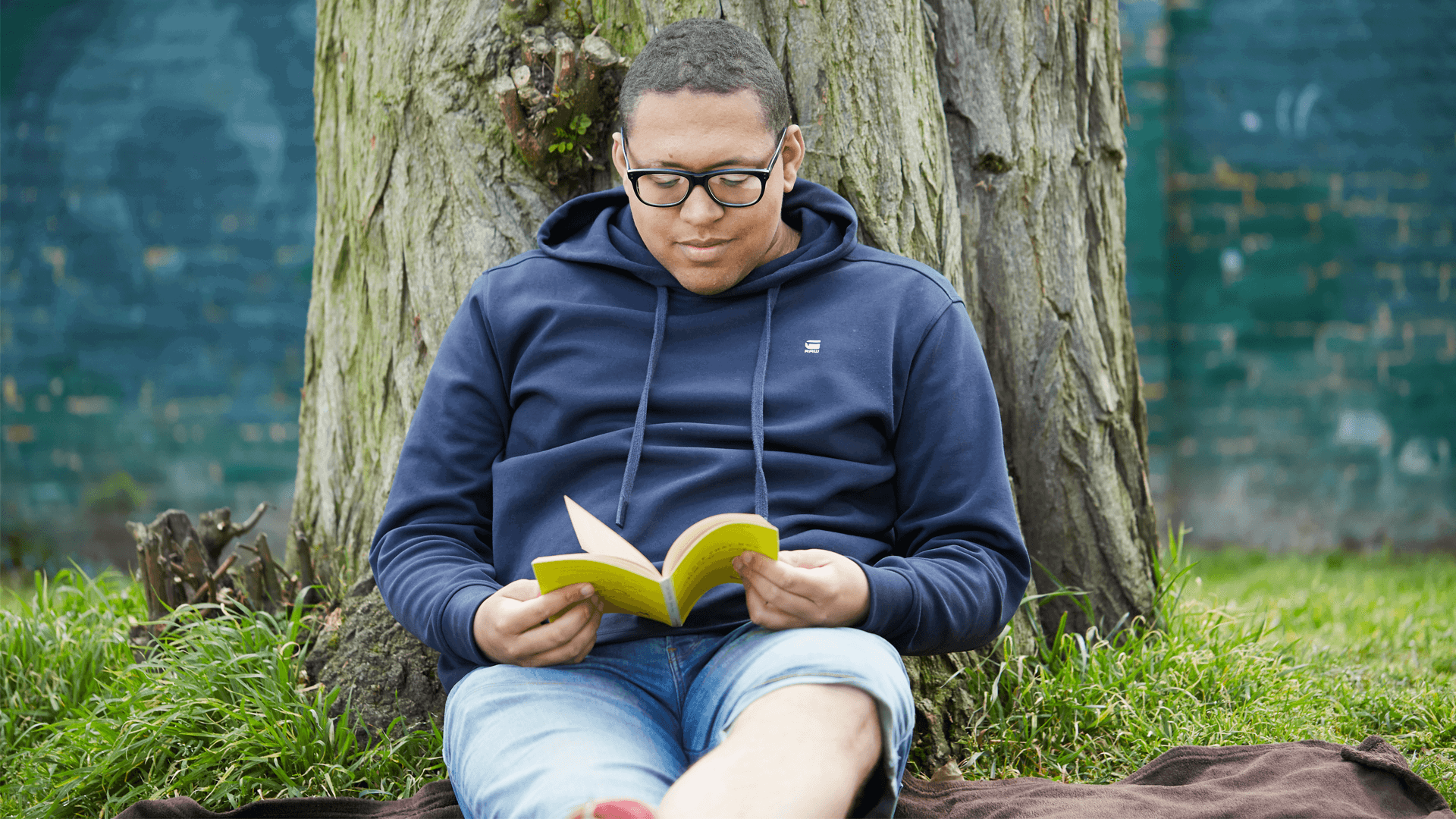 A young man wearing glasses and a navy hoodie. He is sat on the grass and leaning against a tree while reading a book.