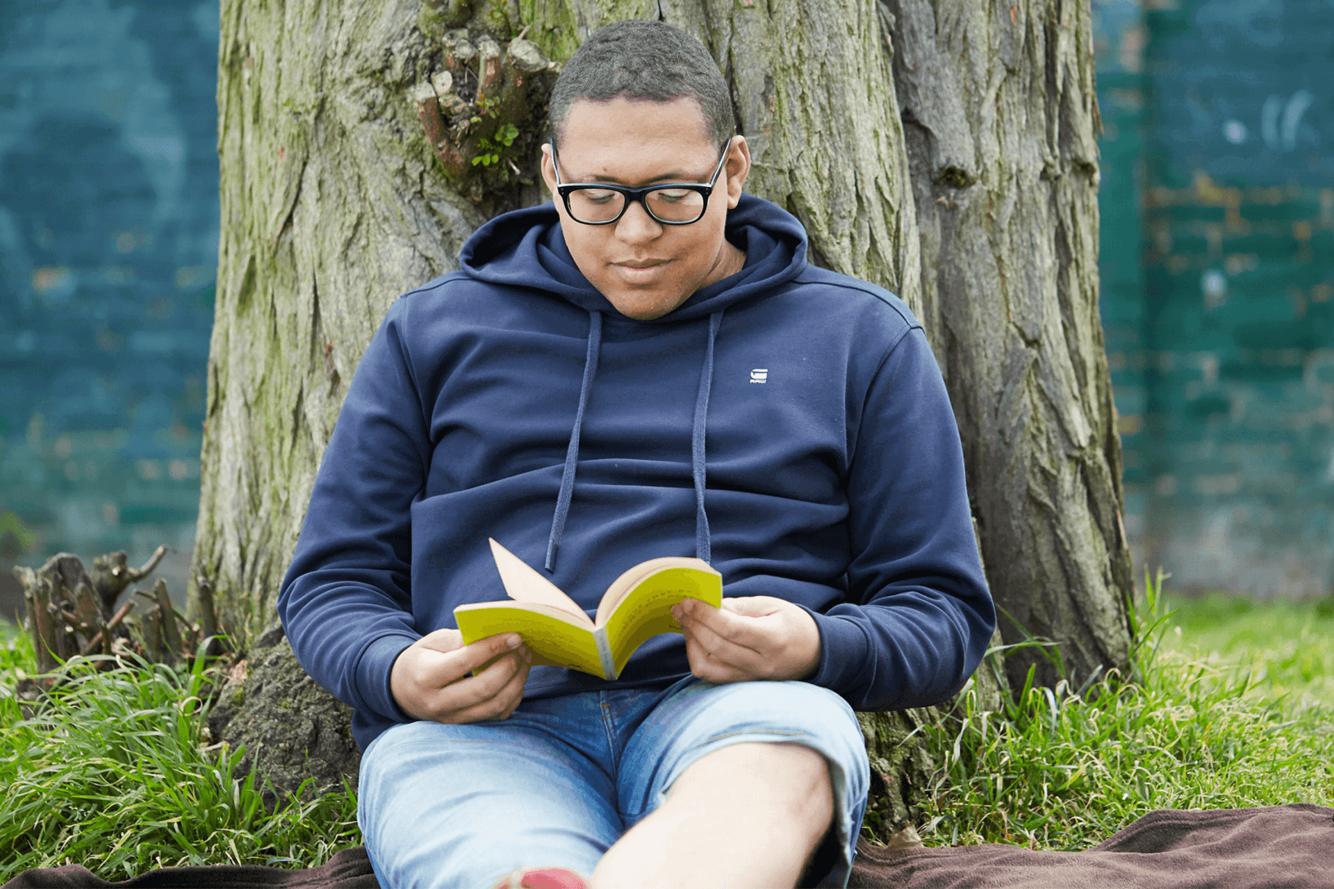 A young man wearing glasses and a navy hoodie. He is sat on the grass and leaning against a tree while reading a book.
