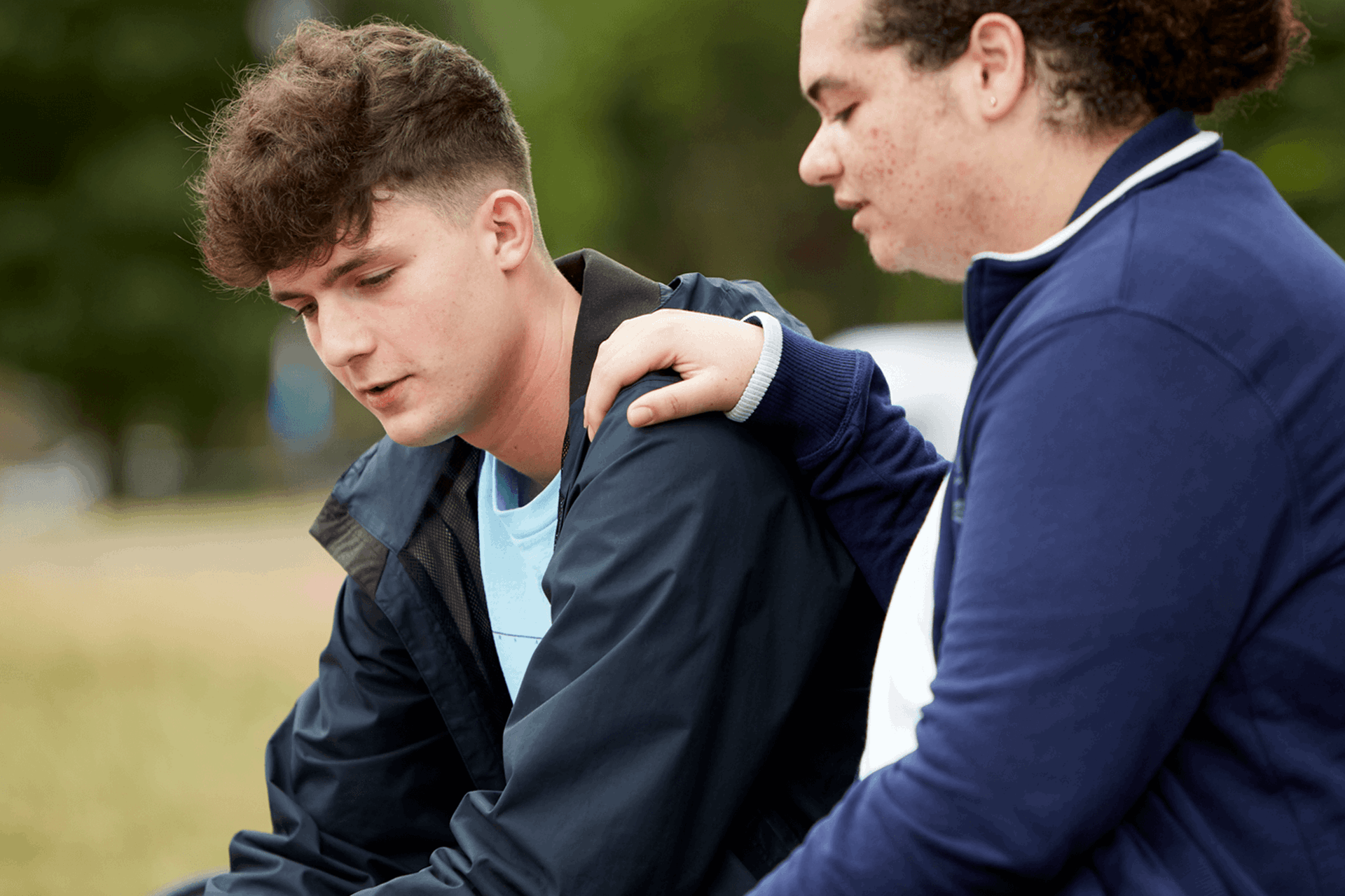 A boy comforting his upset friend by putting with his hand on his shoulder while they sit in a park.