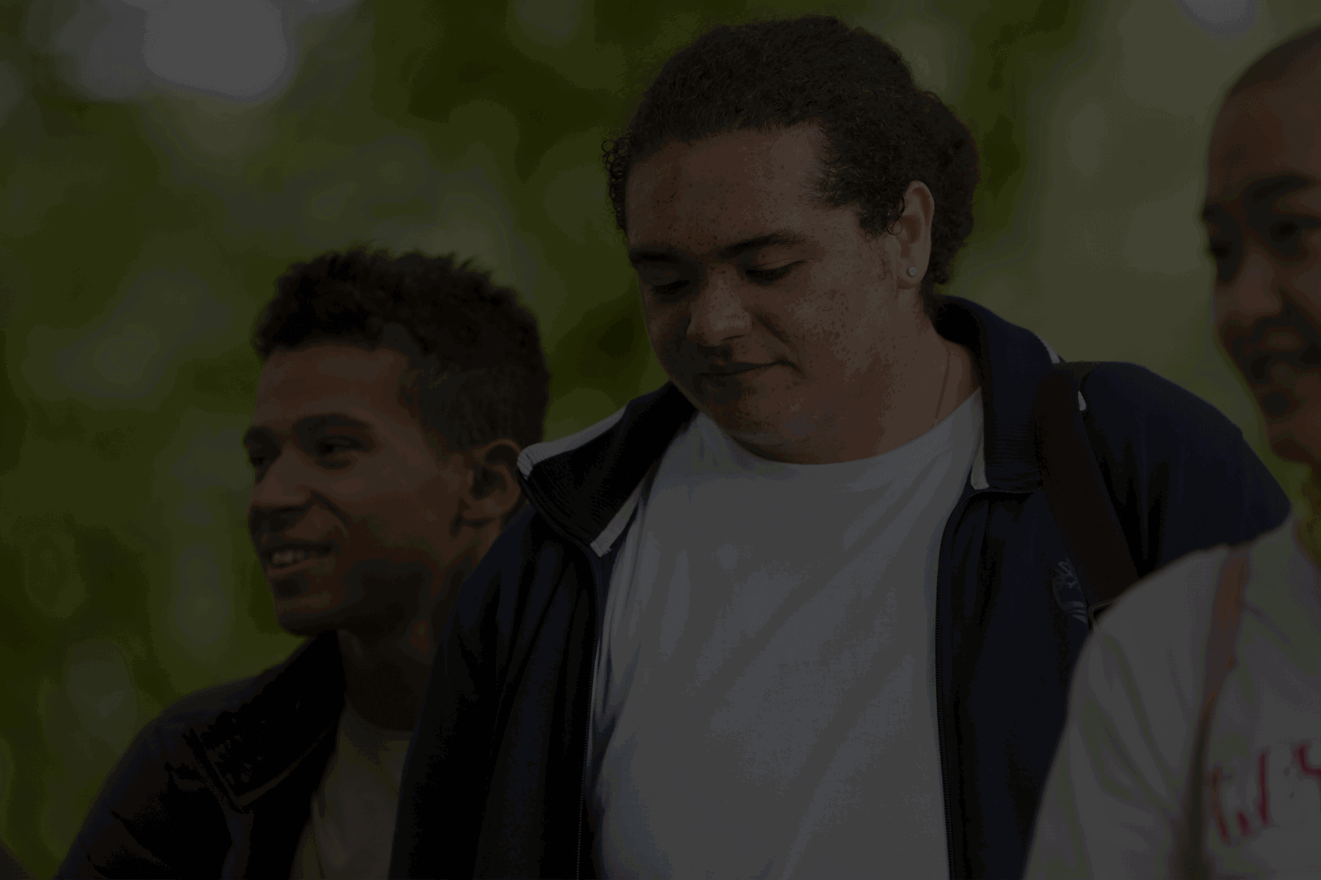 Three young people walk together through a park. On the left the young person is at the back looking to the right. The young person in the middle is looking down. The young person at the front on the right also looks to the right and is wearing yellow headphones.