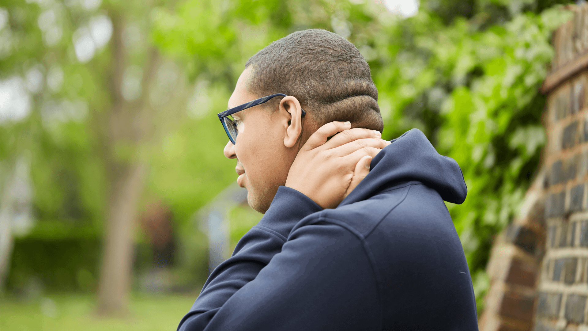 A boy wearing glasses and a black hoodie stands in a park looking worried. He is rubbing the back of his neck with one hand.