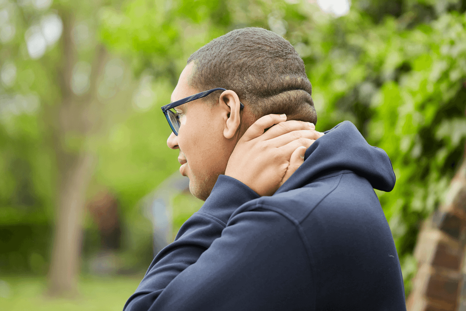 A boy wearing glasses and a black hoodie stands in a park looking worried. He is rubbing the back of his neck with one hand.