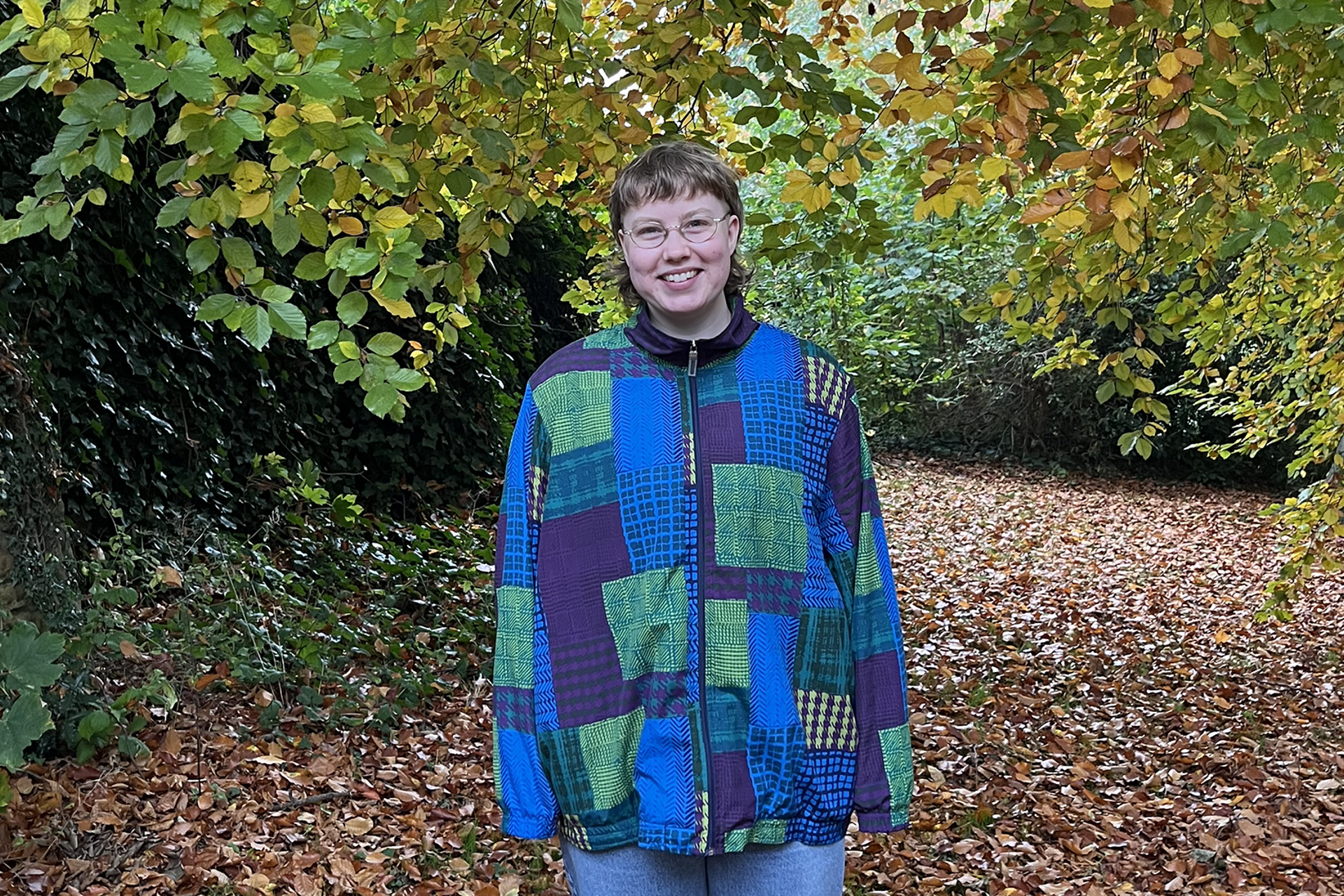 Sofe standing in a forest and smiling. They are wearing glasses and a blue and green patterned jacket.