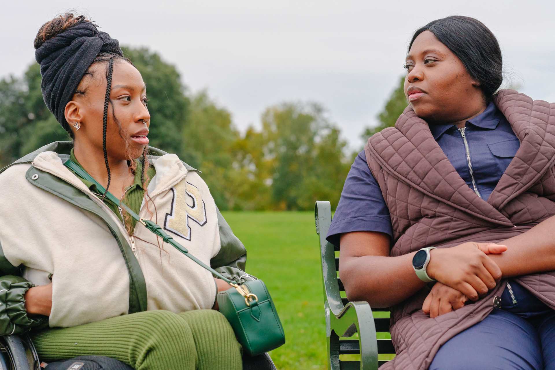 A young Black woman in a wheelchair talking to an older Black woman on a bench in the park.