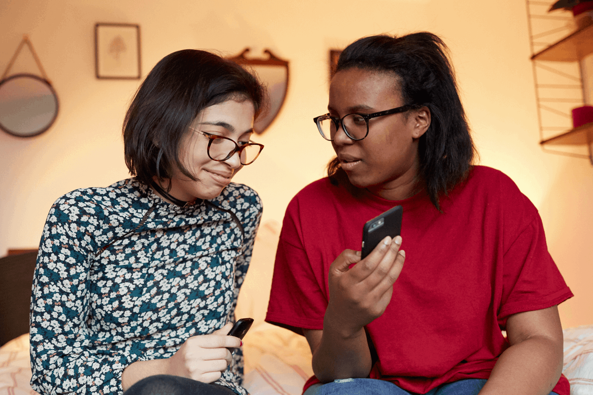 Two girls sitting in a bedroom. One is showing the other something on her phone.