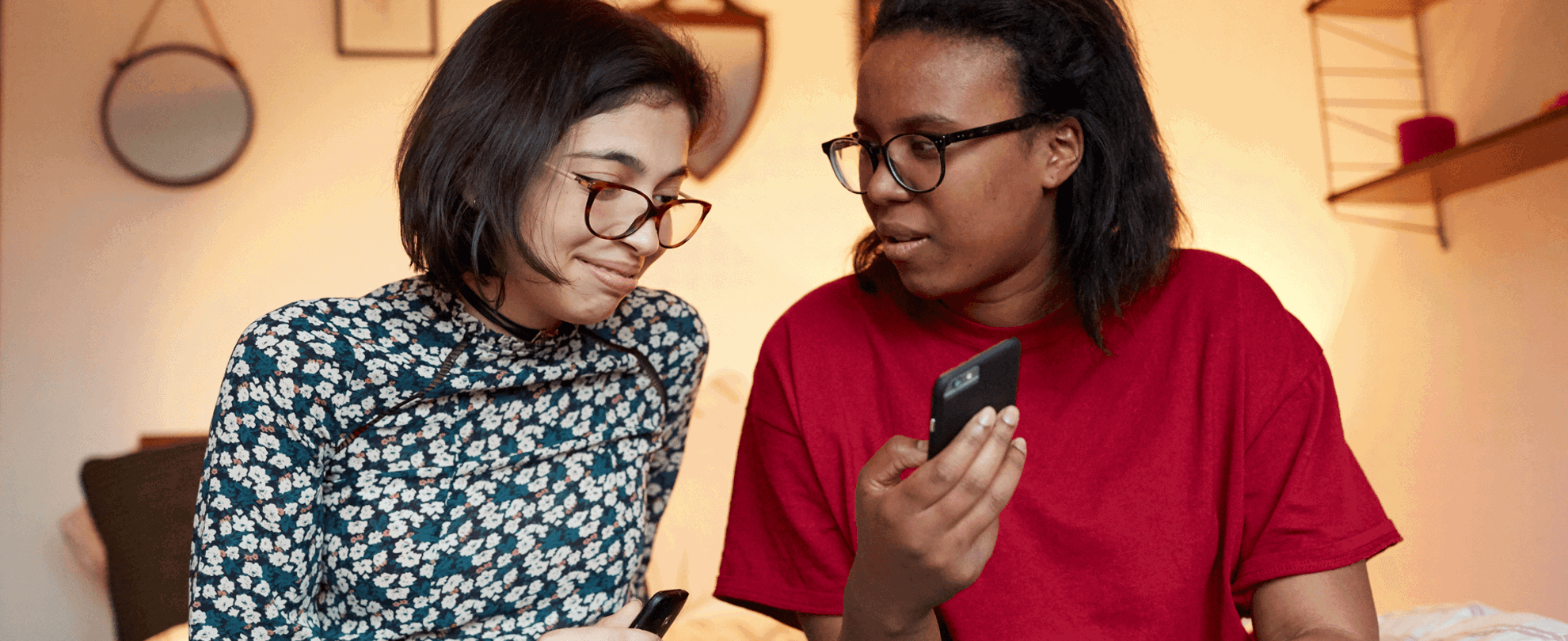 Two girls sitting in a bedroom. One is showing the other something on her phone.