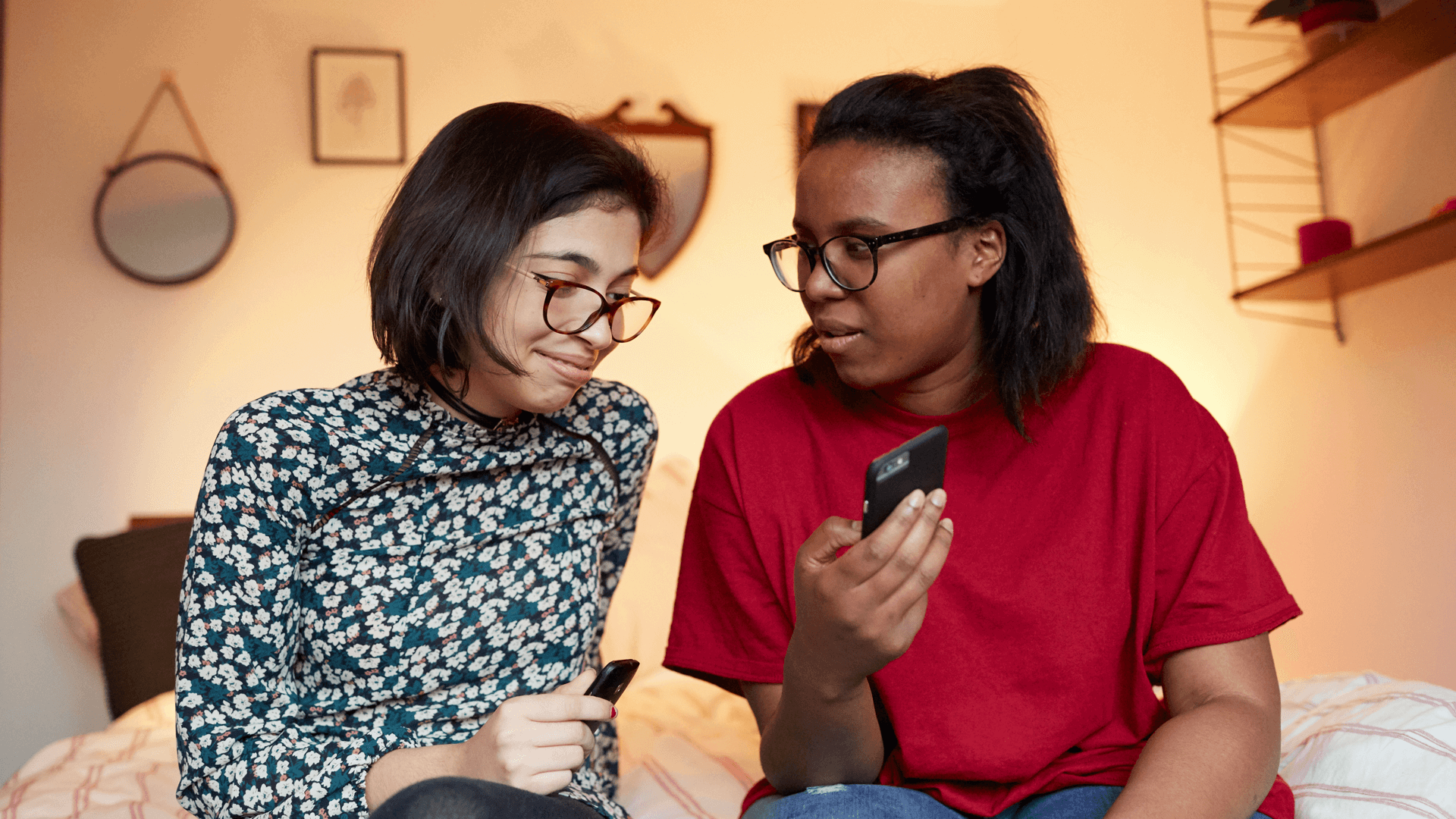 Two girls sitting in a bedroom. One is showing the other something on her phone.