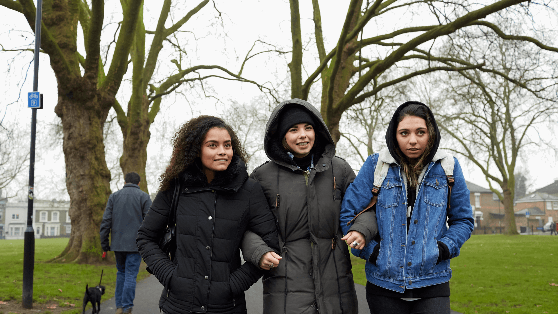 wide shot of three girls walking with linked arms on a tree lined street on a chilly day