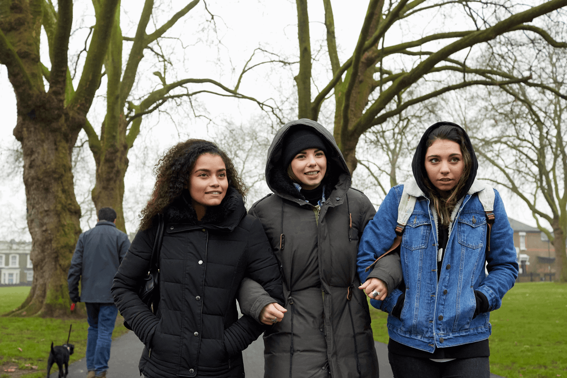 wide shot of three girls walking with linked arms on a tree lined street on a chilly day